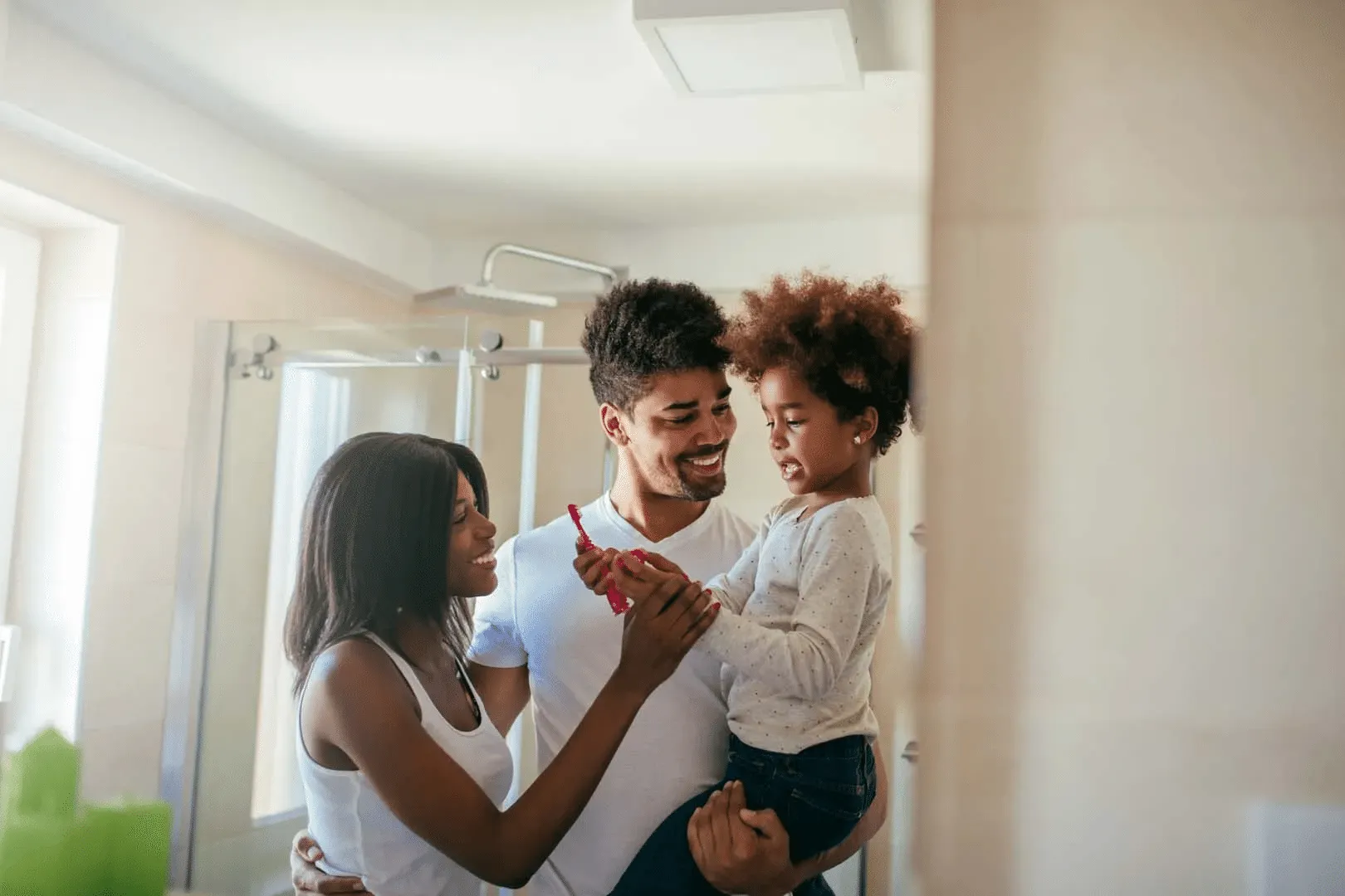Smiling parents helping their child hold a toothbrush in a bright bathroom.