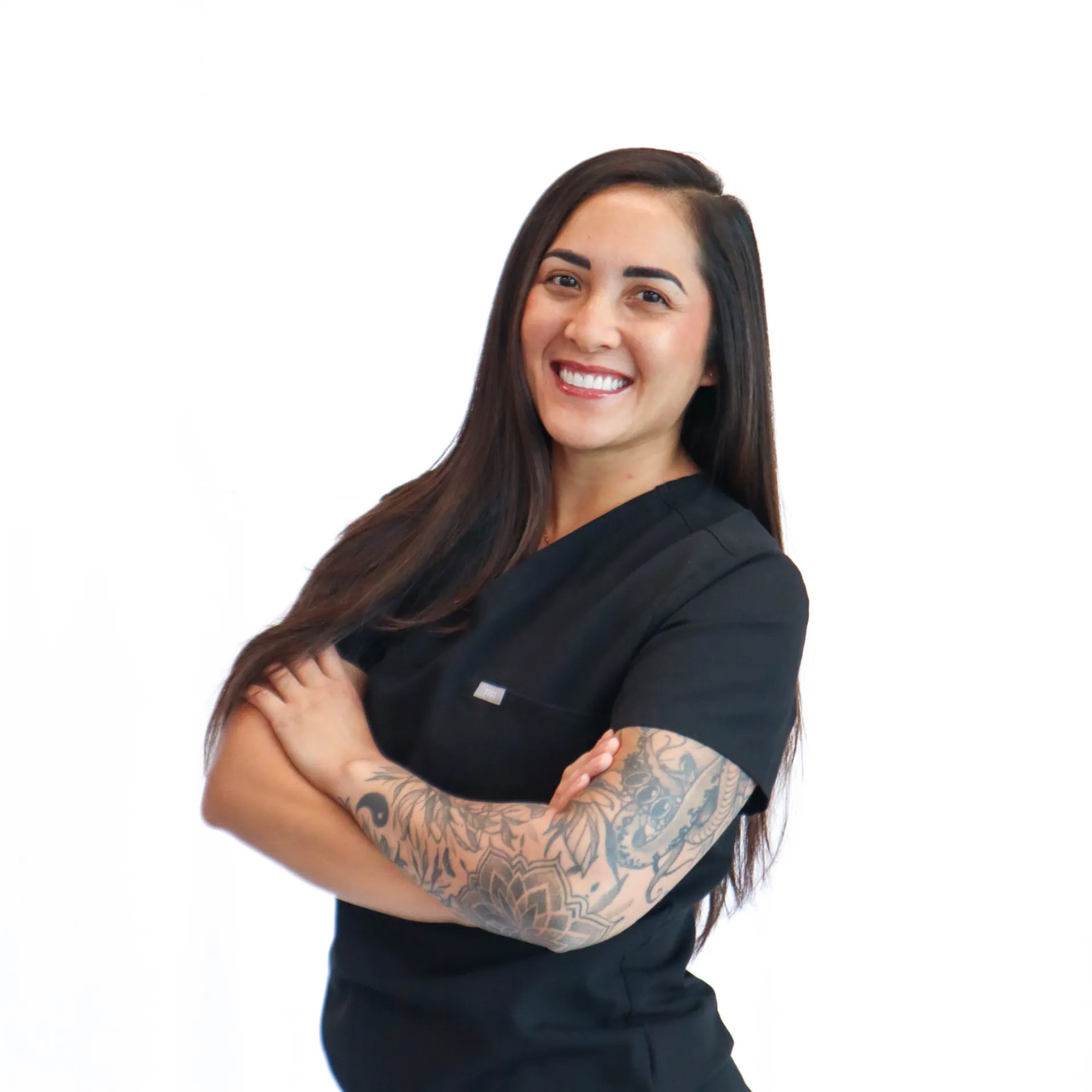 Smiling woman with long dark hair wearing black medical scrubs and showing a detailed tattoo sleeve on her left arm.
