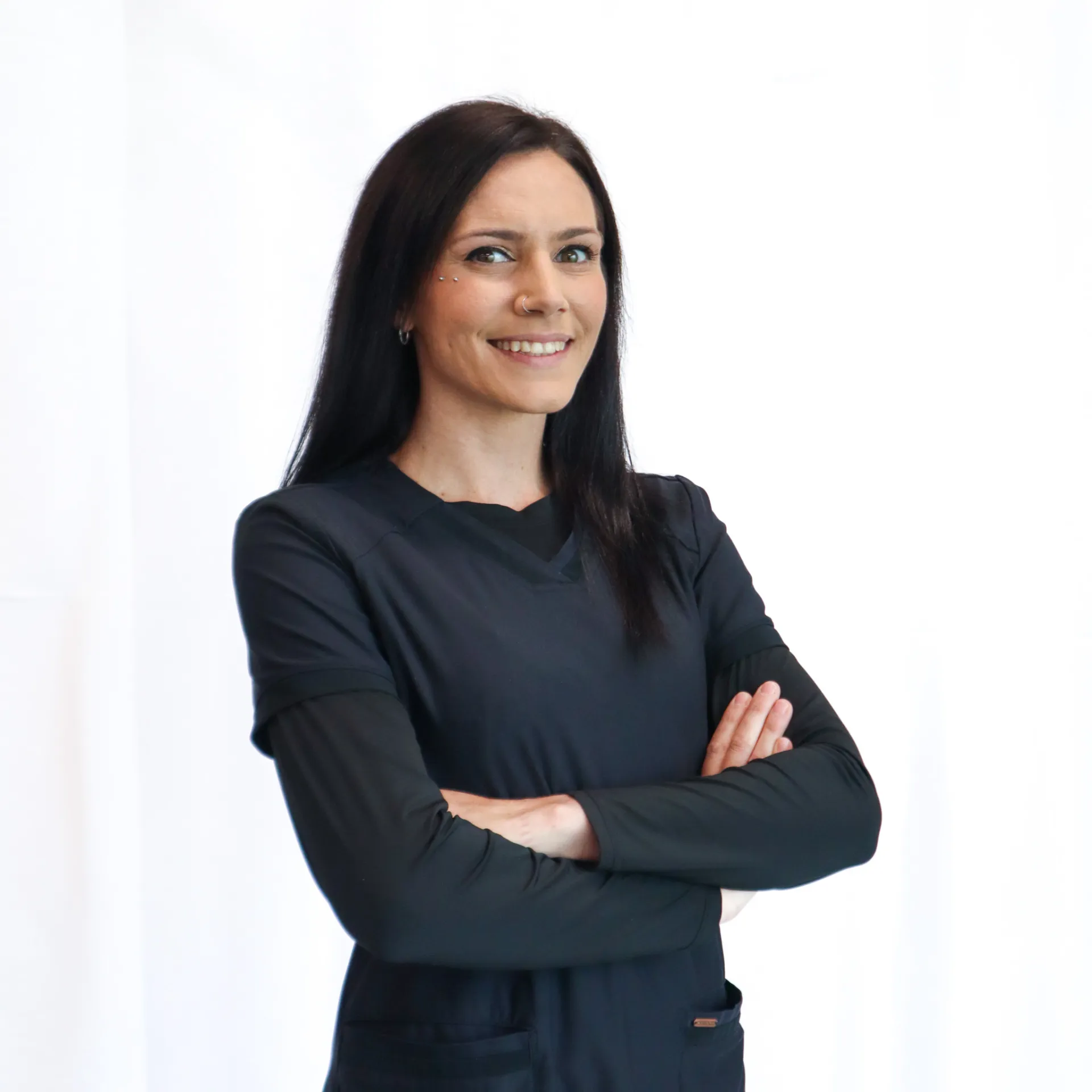 Smiling woman with long dark hair and piercings wearing black scrubs with arms crossed against white background.
