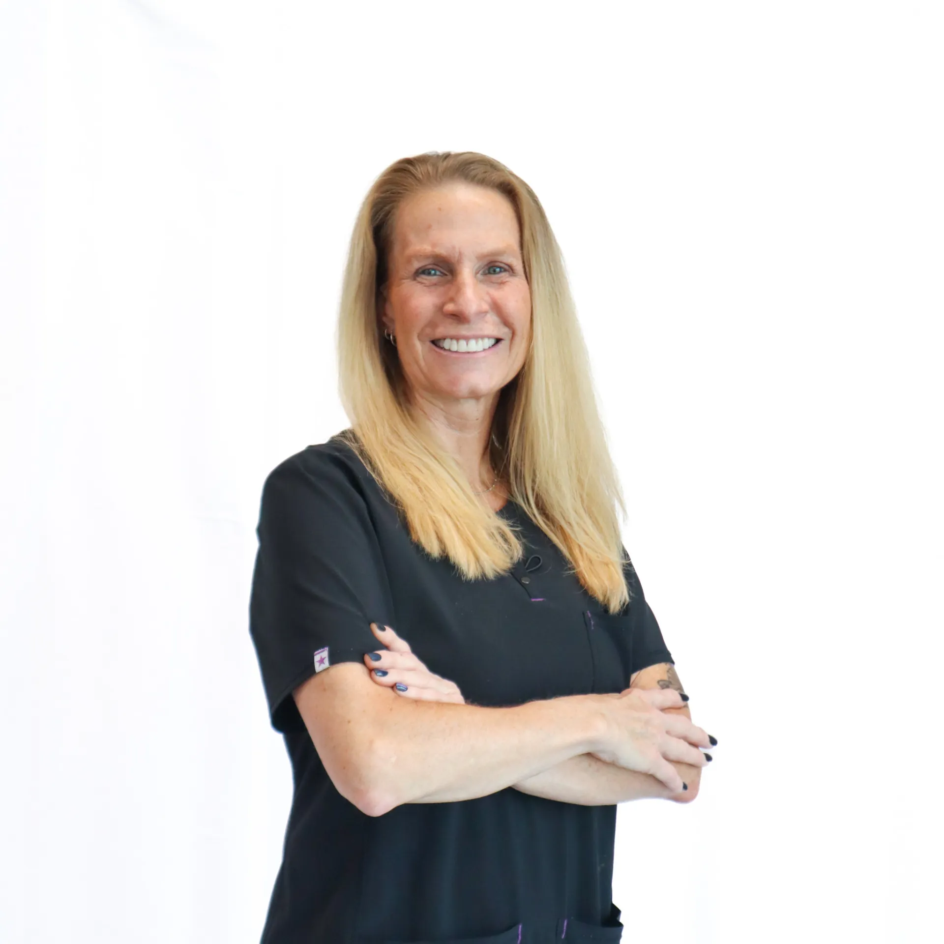 Woman with long blonde hair in black scrubs smiling with arms crossed against white background.