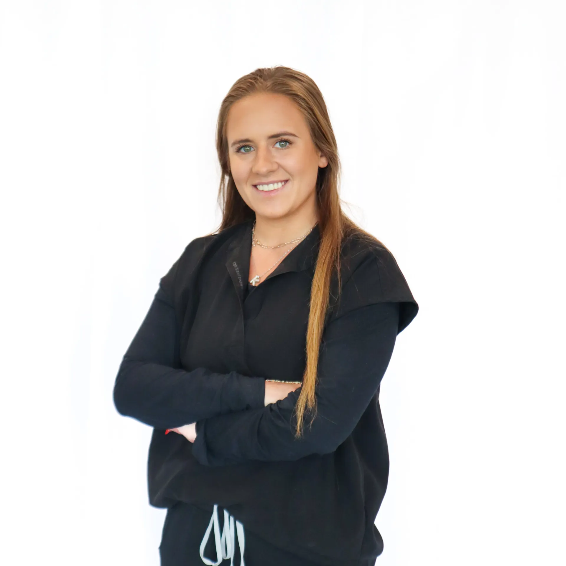 Smiling young woman with long brown hair wearing black clothing and standing with arms crossed against a white background.