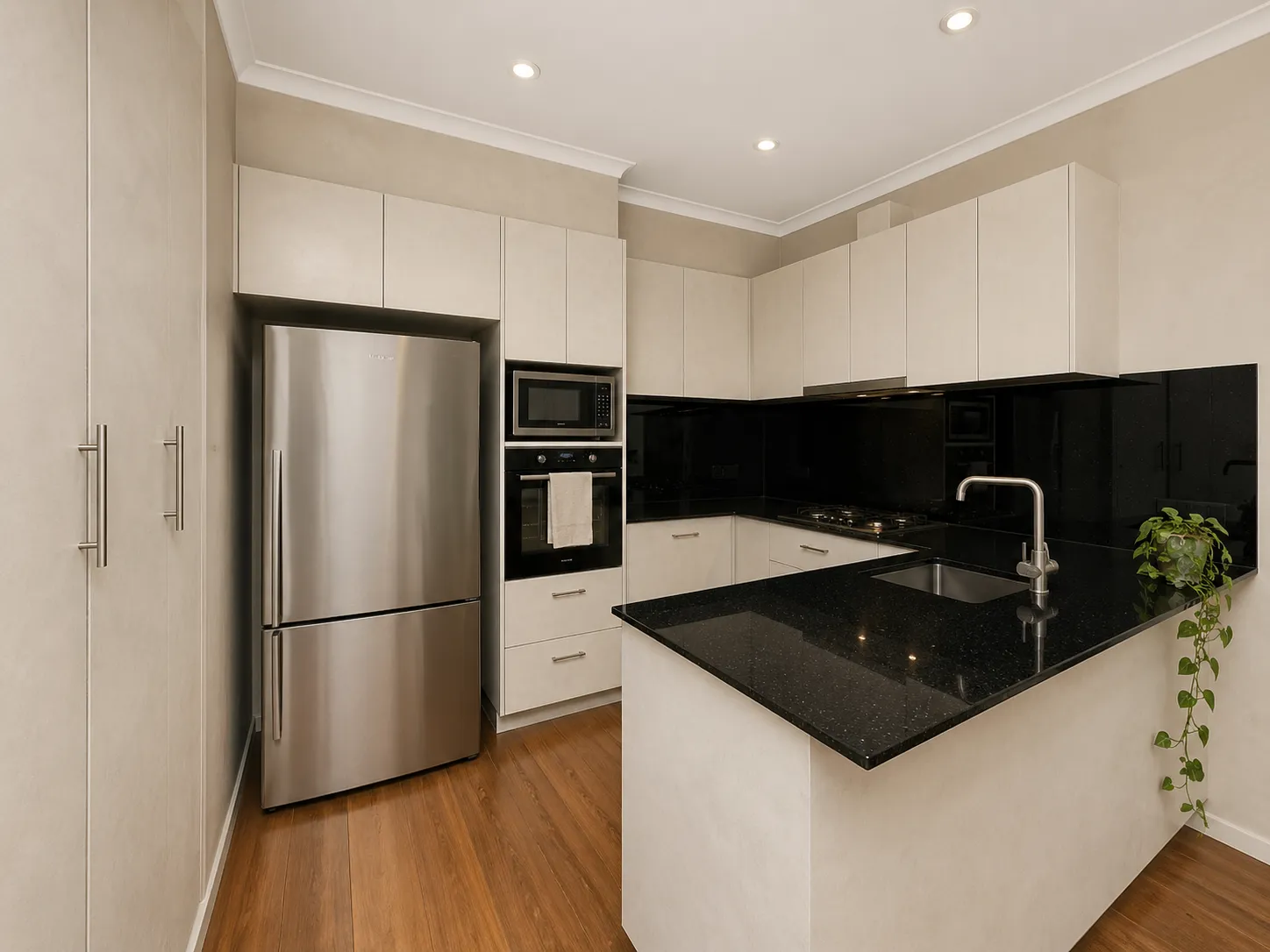 Modern kitchen with beige cabinets, black granite countertops, stainless steel refrigerator, built-in oven, microwave, gas stove, and a small green plant on the counter.