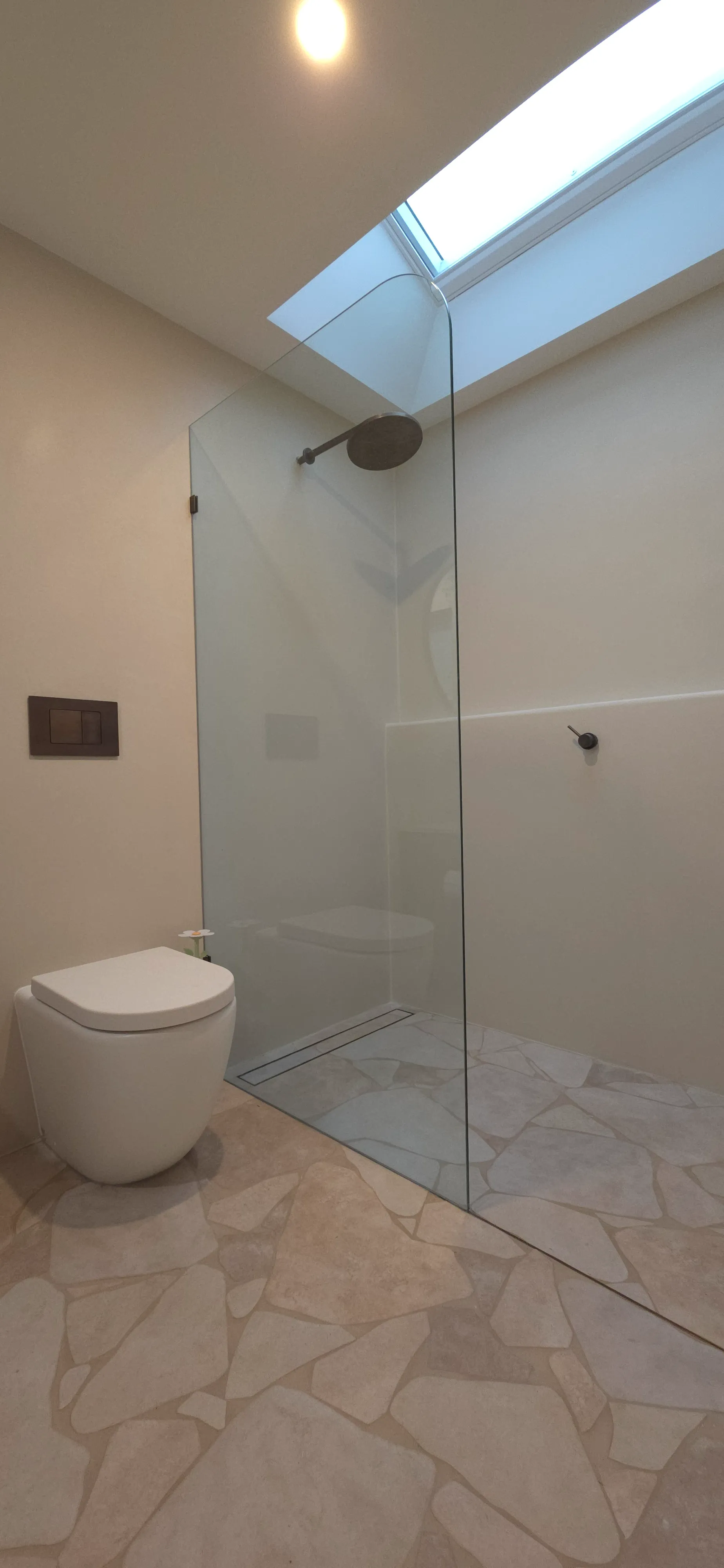 Modern minimalistic bathroom with a wall-mounted toilet, a walk-in shower with glass partition, and skylight above.