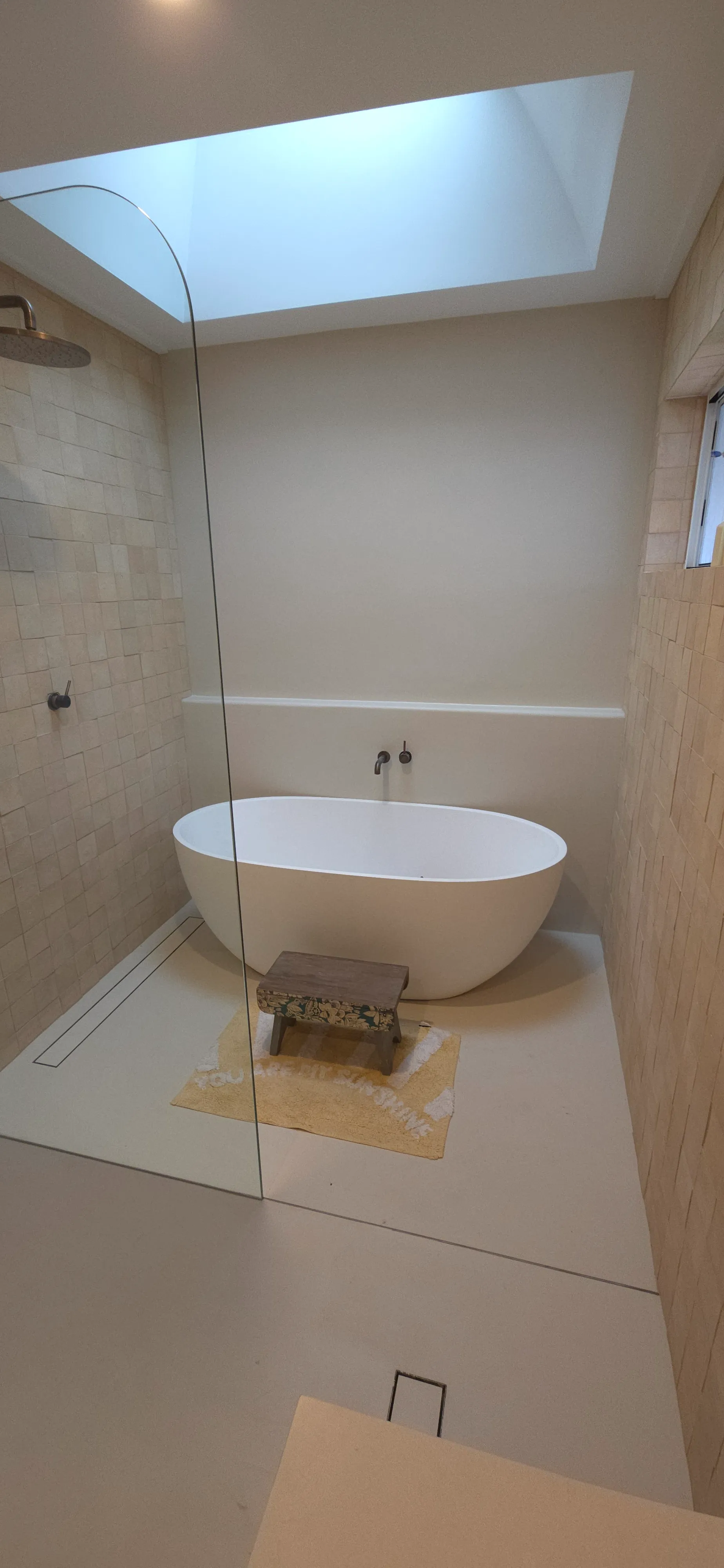 Modern bathroom with a white freestanding bathtub, small wooden stool on a yellow bath mat, beige tiled walls, and a skylight above.