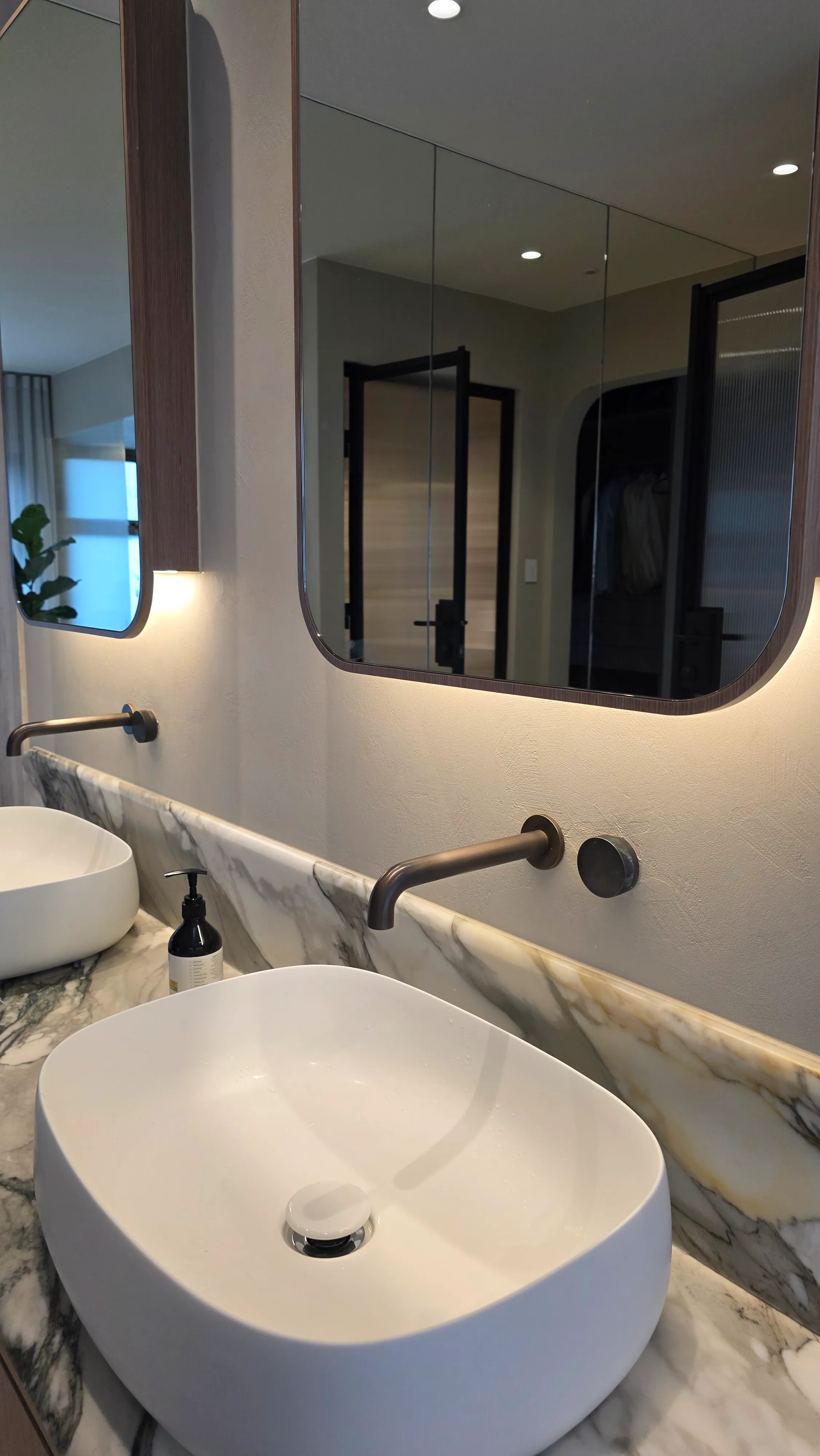 Modern bathroom countertop with white vessel sinks, wall-mounted bronze faucets, and backlit mirrors above a marble surface.