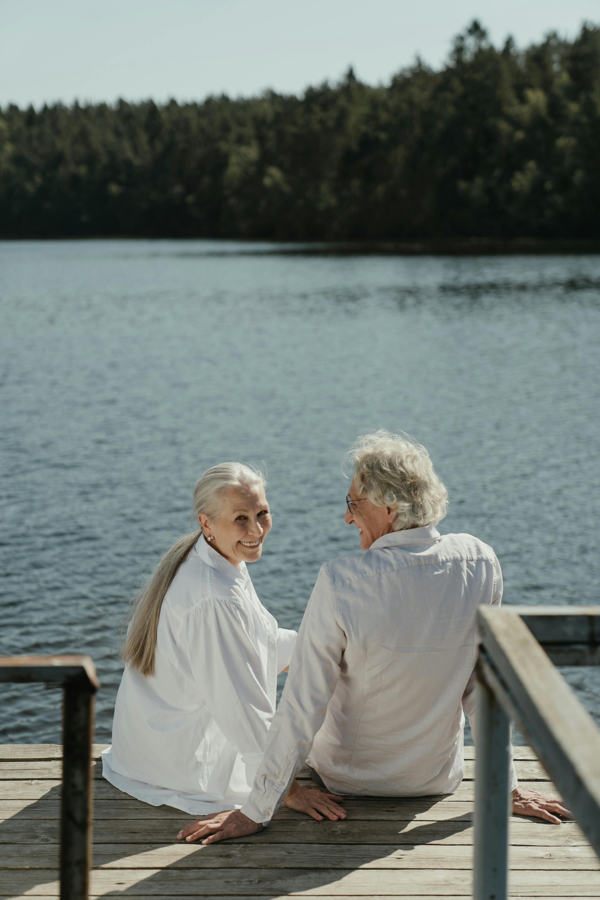 Elderly couple sitting on a wooden dock by a lake, enjoying a sunny day together.