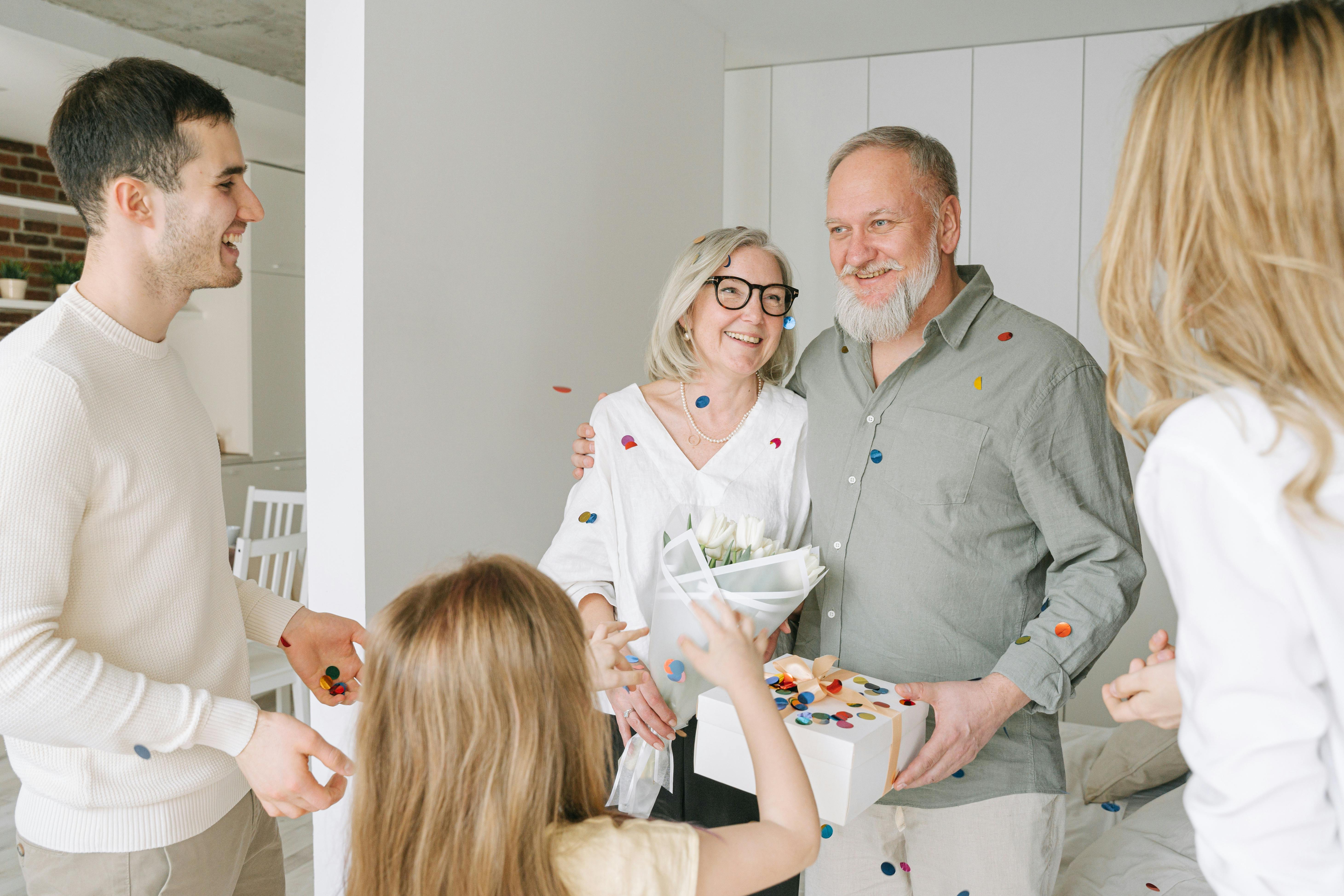 Smiling elderly couple with flowers and gift surrounded by family celebrating indoors with confetti.