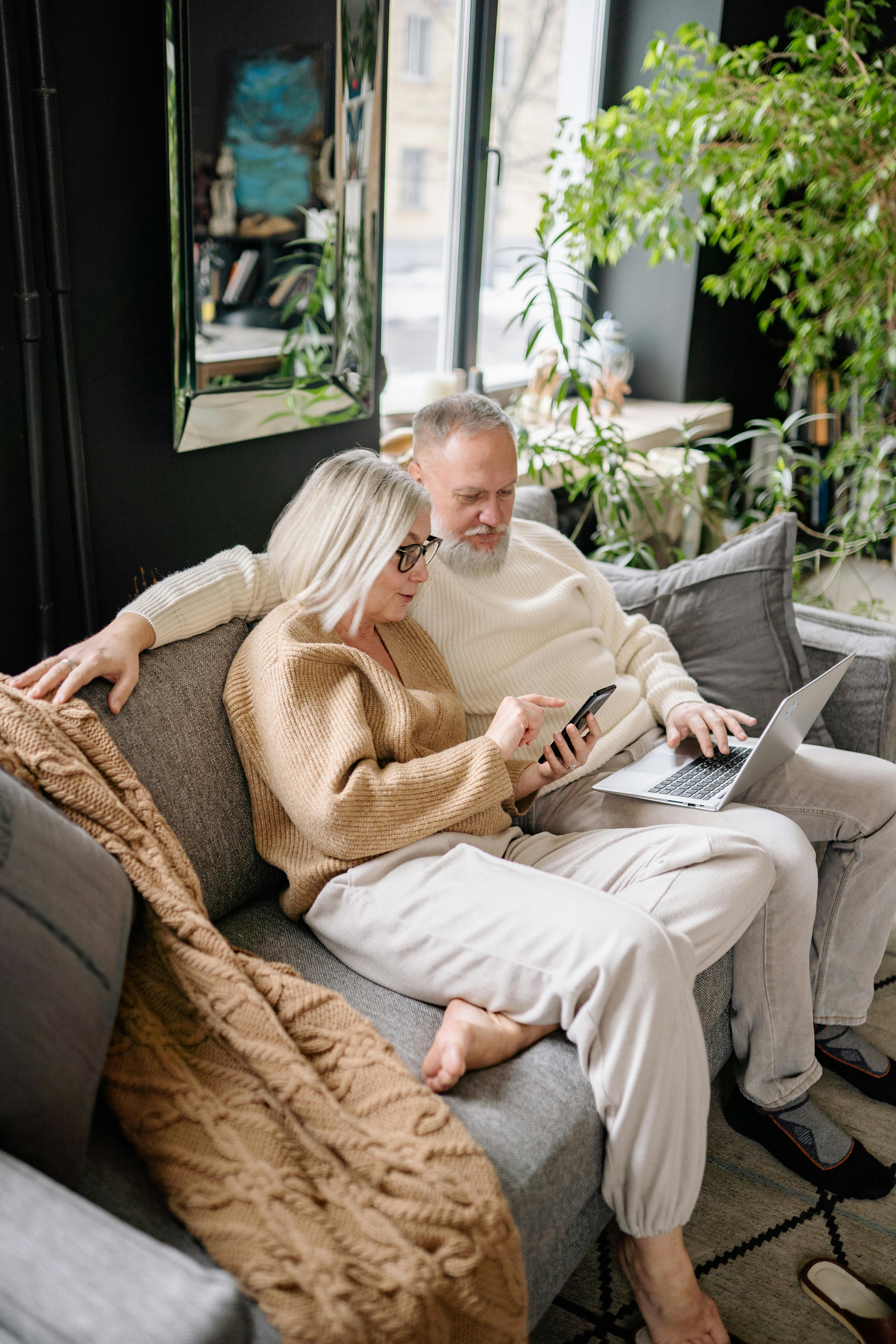 Mature couple sitting on a couch using a phone and laptop together in a cozy, plant-filled room.