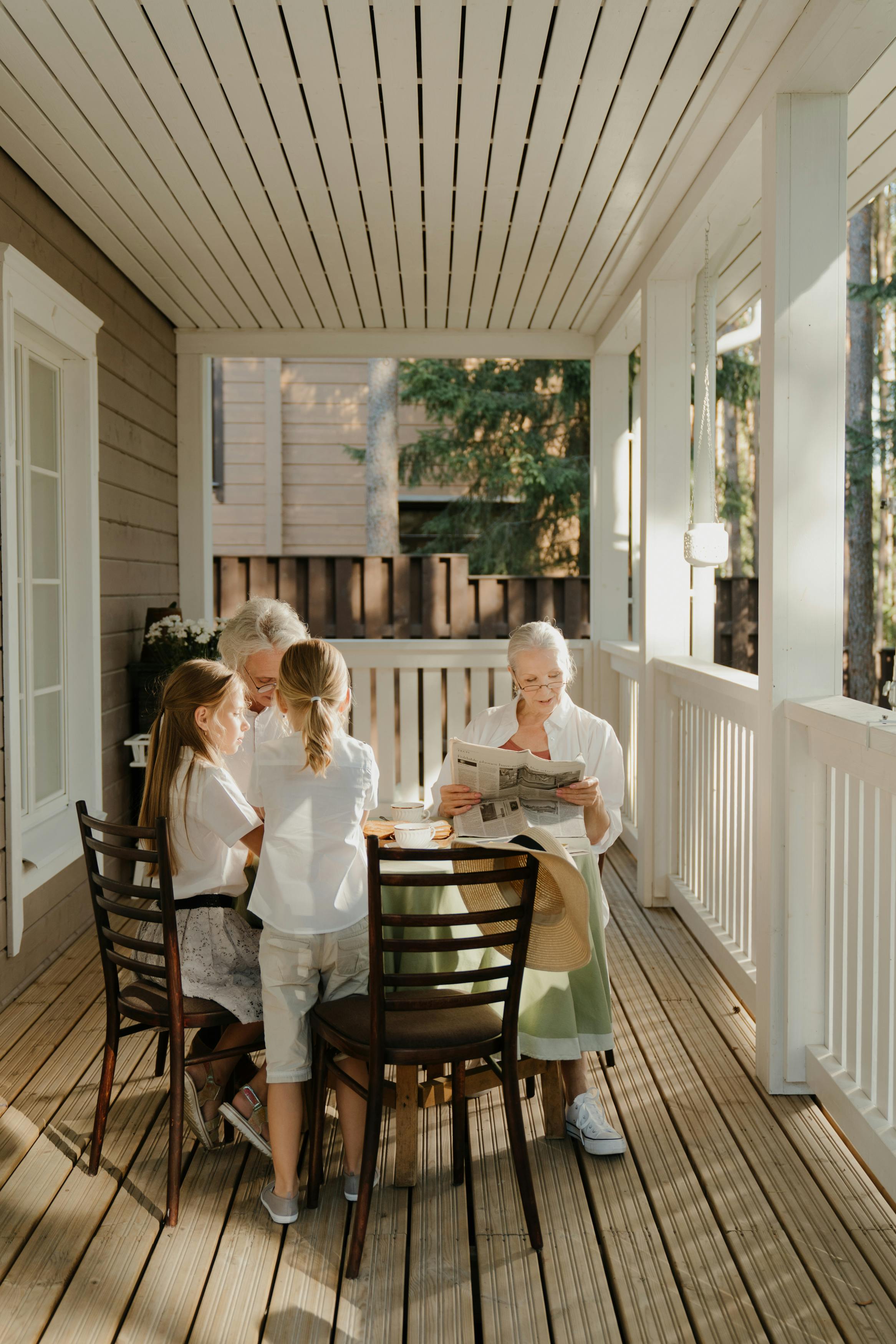 Two elderly women and two young girls sitting and standing around a table on a sunlit porch.