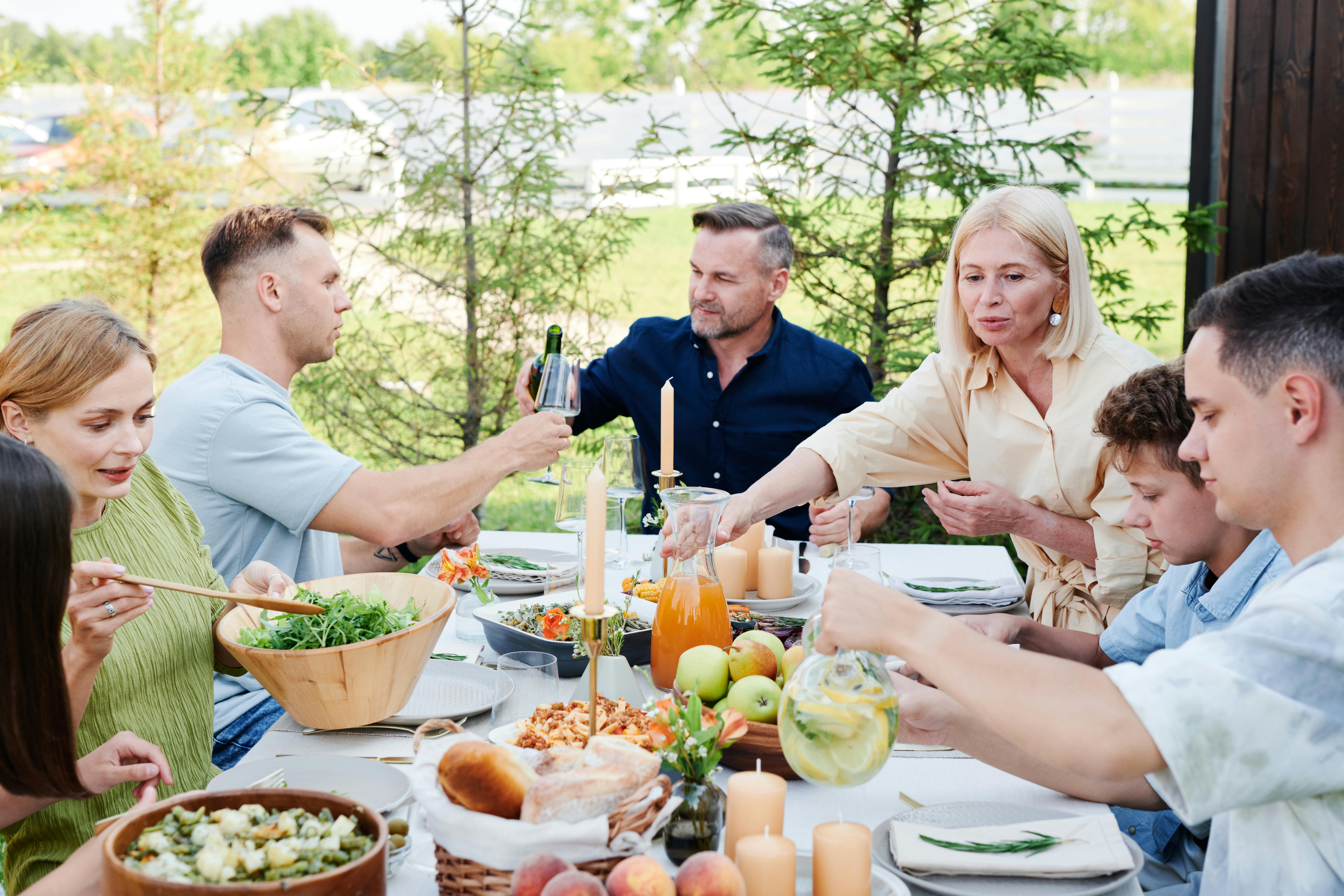 Family enjoying outdoor meal with salad, drinks, bread, fruit, and candles on a sunny day.