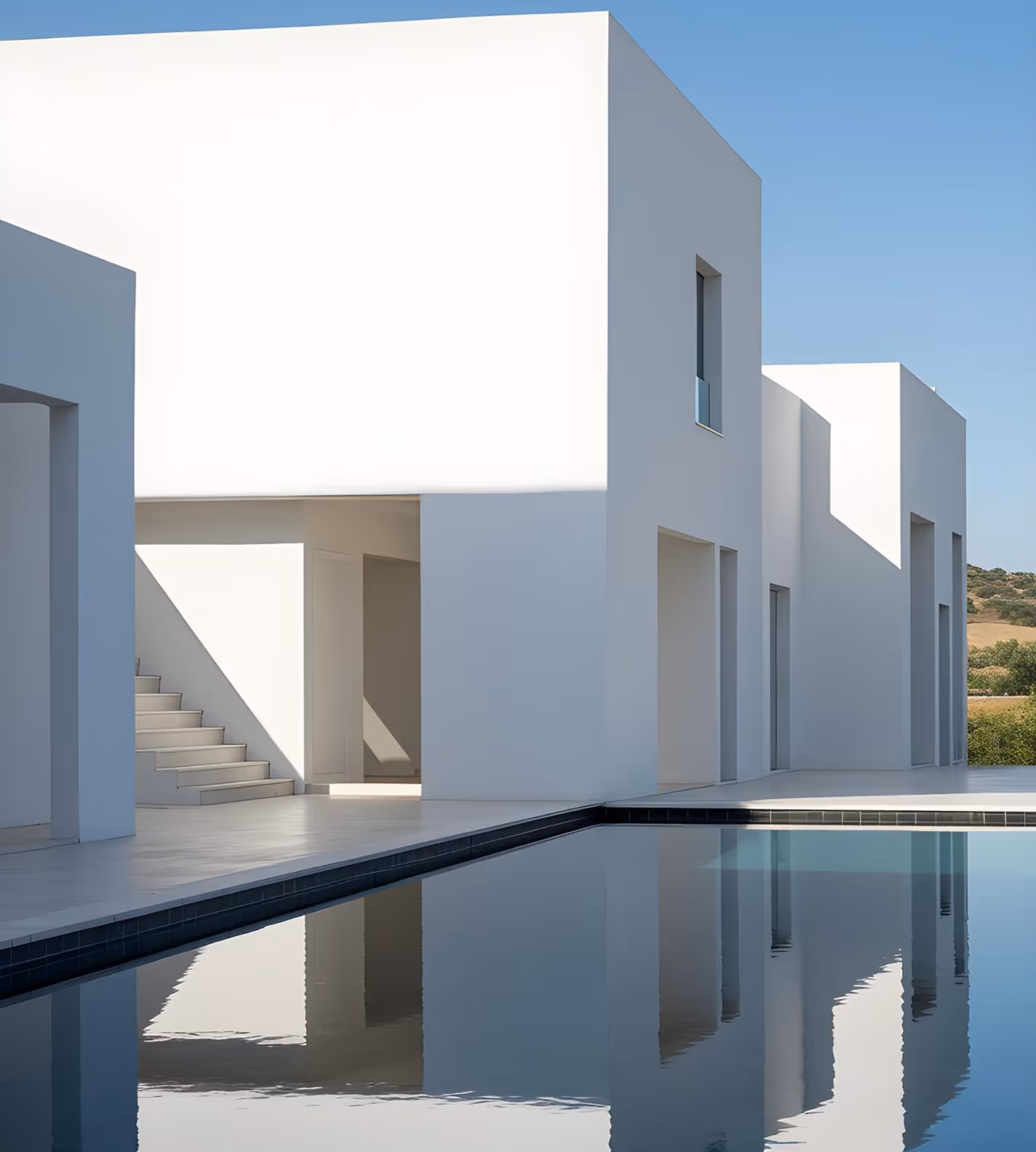 Modern white cubic building with large windows and a calm reflecting pool in front under a clear blue sky.