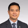 Portrait of a smiling Asian man wearing a dark suit, white shirt, and striped tie against a gray background.