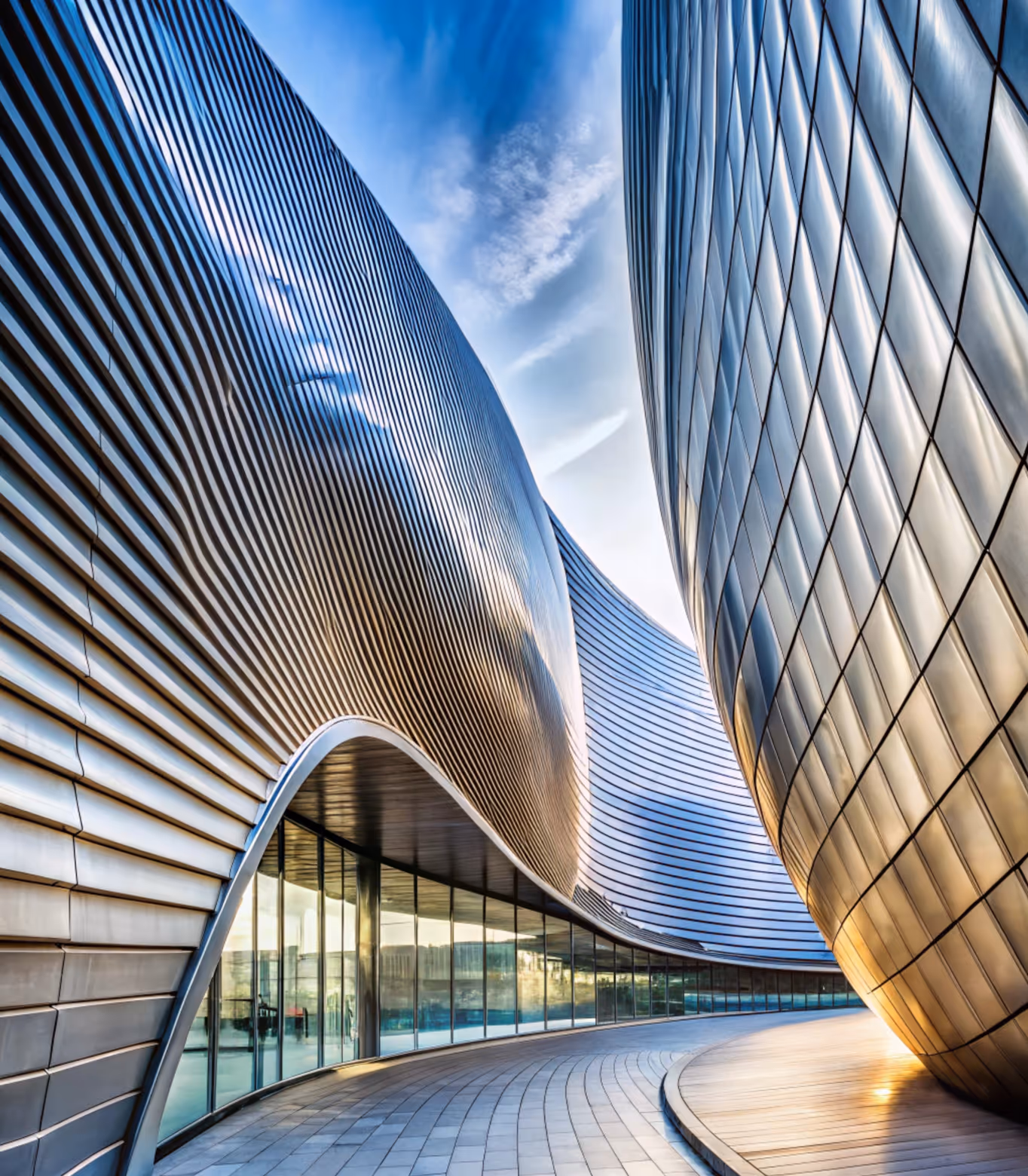 Curved modern building with reflective metal panels and glass windows under a blue sky with clouds.