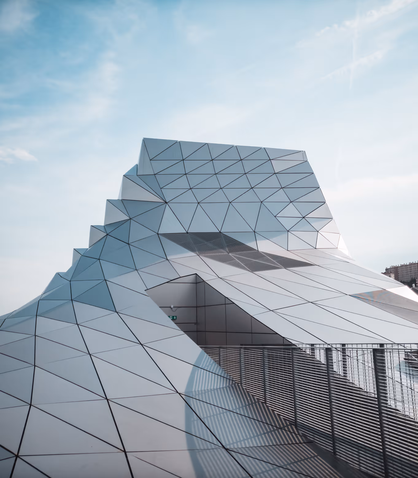Modern architectural structure with angular, triangular glass panels and a walkway with metal railings.