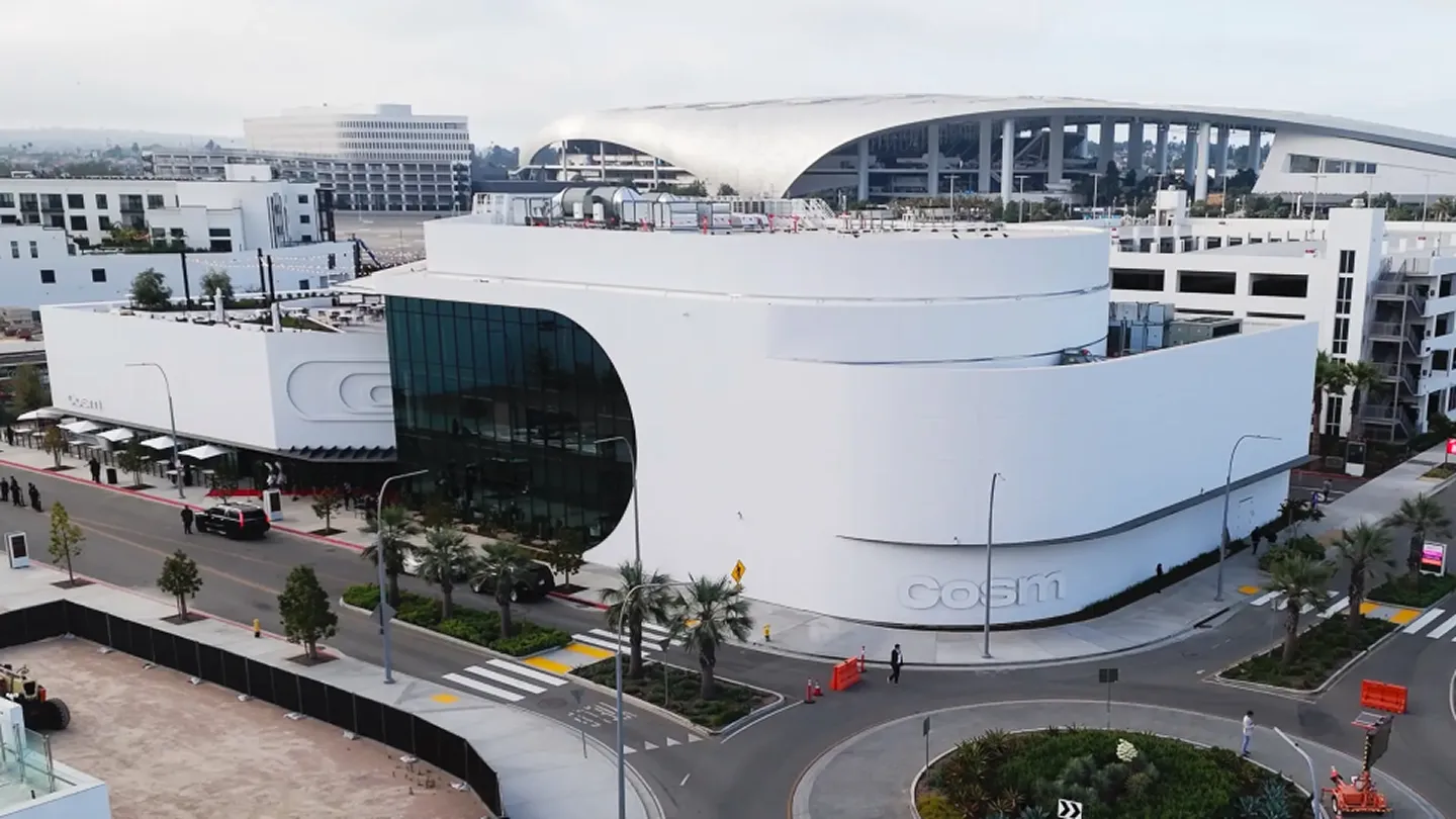 Aerial view of the modern white building Cosm with large curved glass windows, surrounded by roads and palm trees, with a stadium visible in the background.