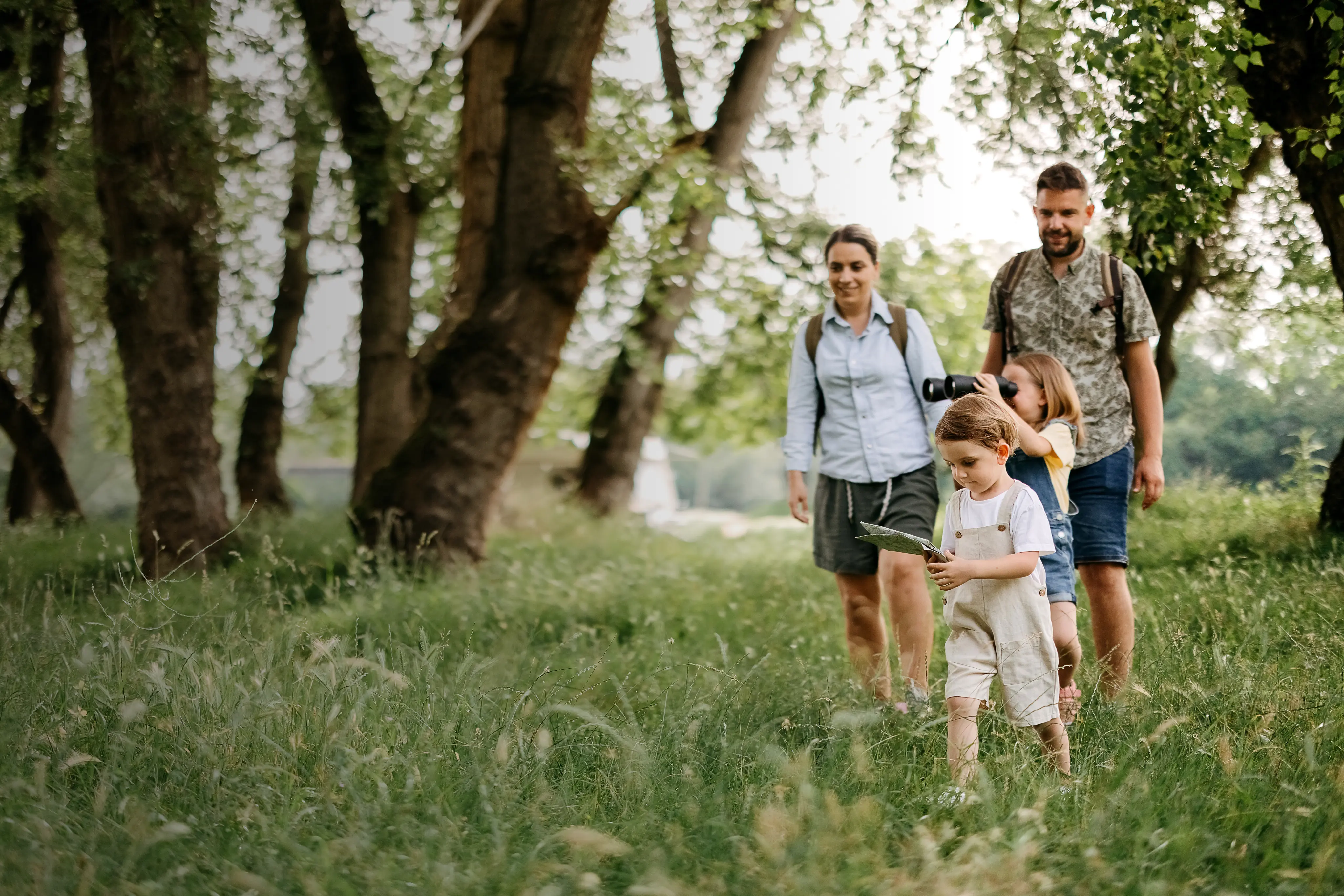 A family of four walking through a grassy forest path, with a young boy holding leaves and a girl looking through binoculars.
