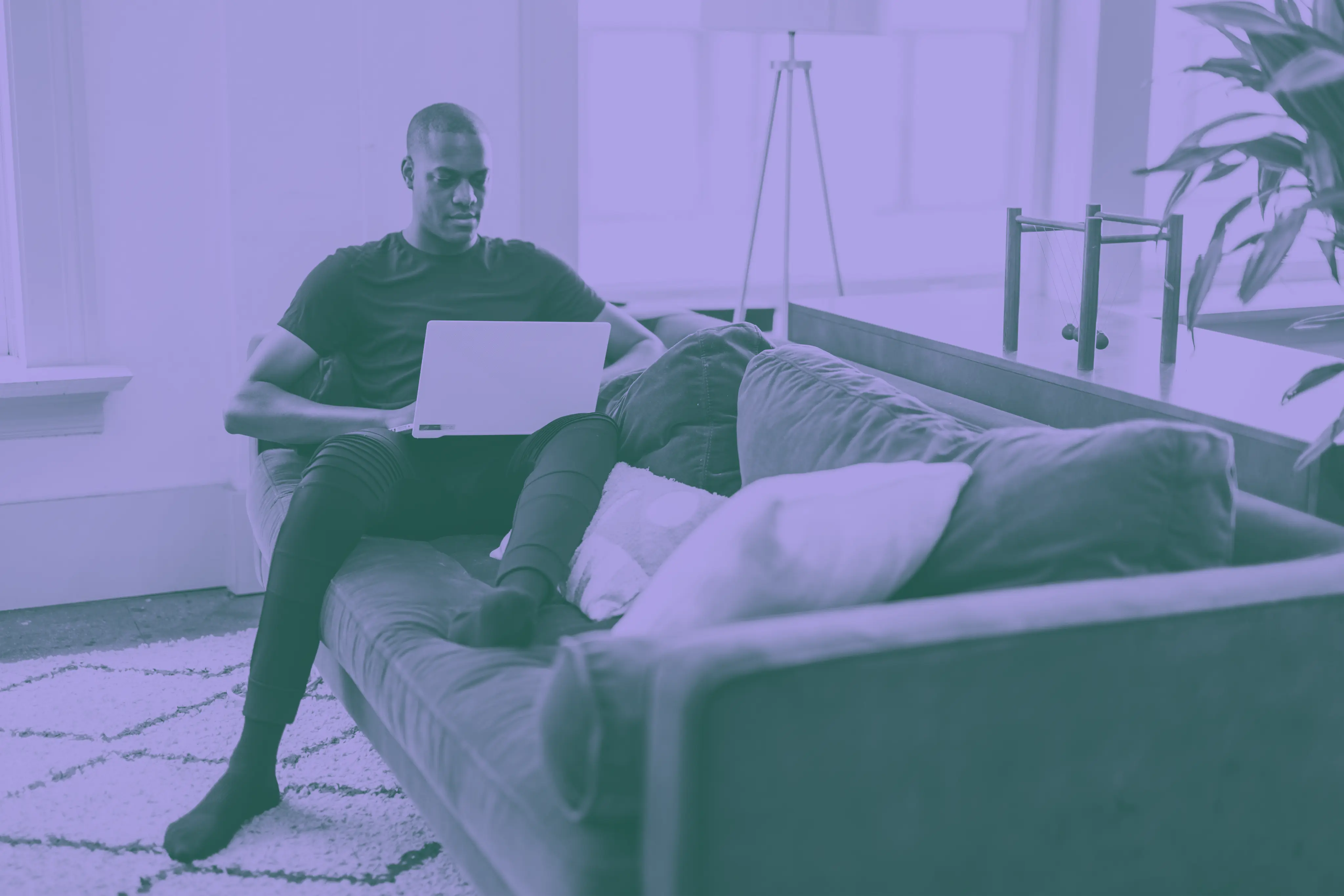 Man sitting on a couch working on a laptop in a bright living room.