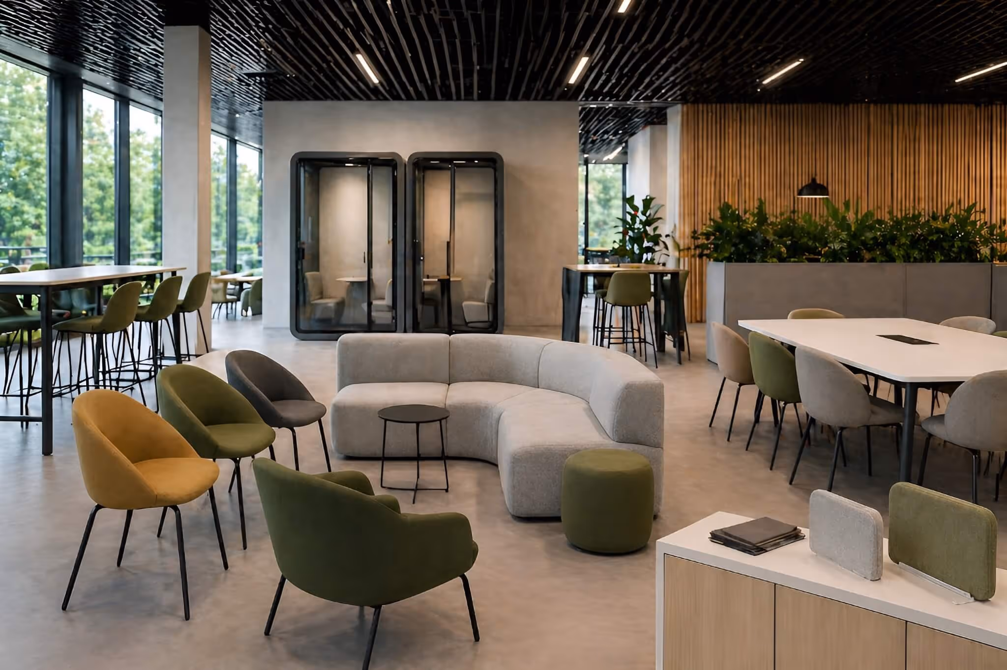 Modern office lounge with curved gray sofa, colorful chairs, small tables, and glass phone booths.