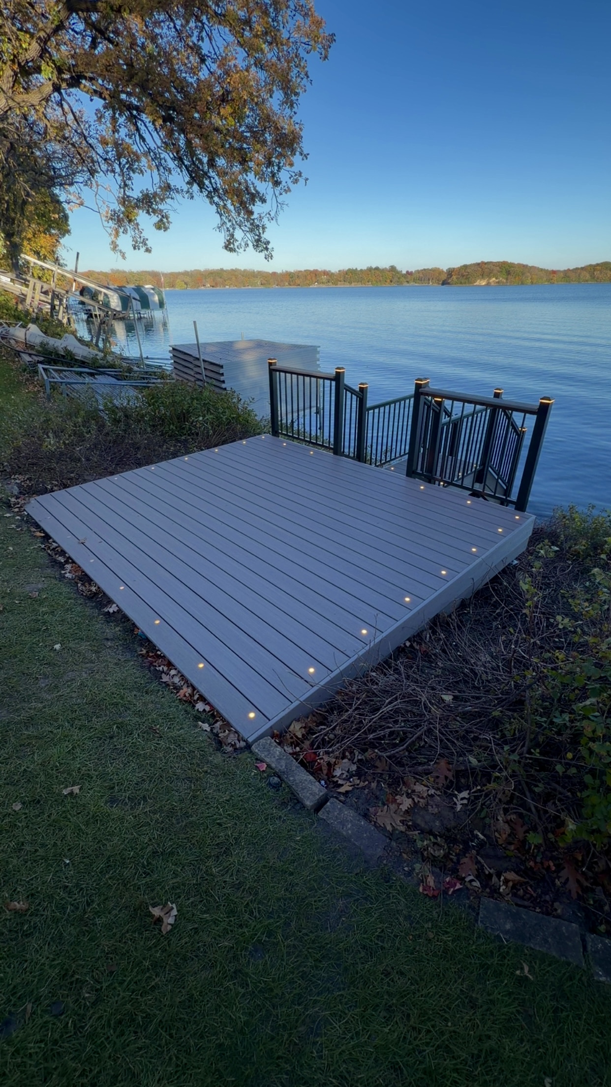 A wooden deck with built-in lights overlooking a calm lake with a clear blue sky and autumn trees in the background.