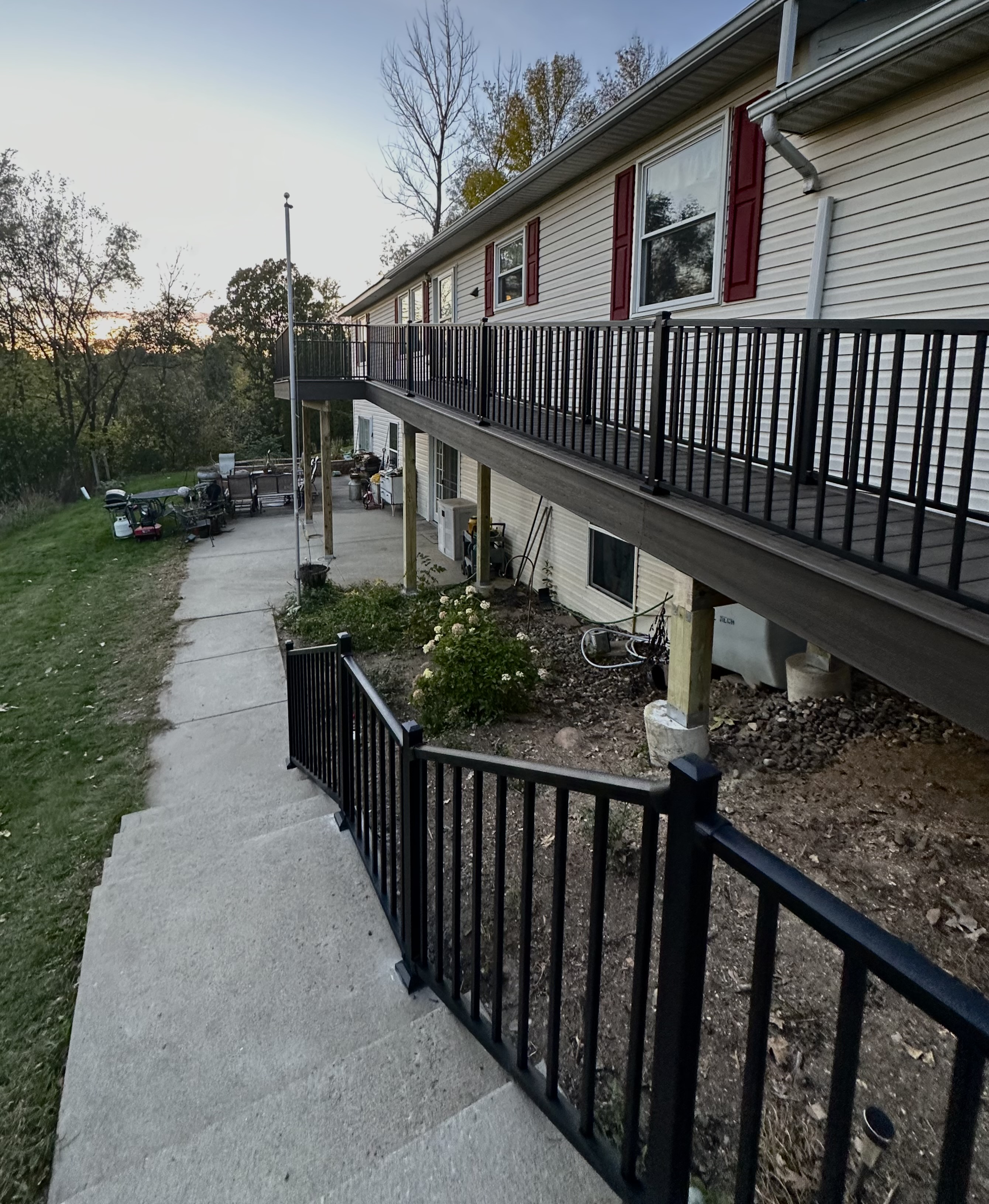 Concrete stairs with black metal railing leading to a backyard patio under a raised deck attached to a beige house with red window shutters.