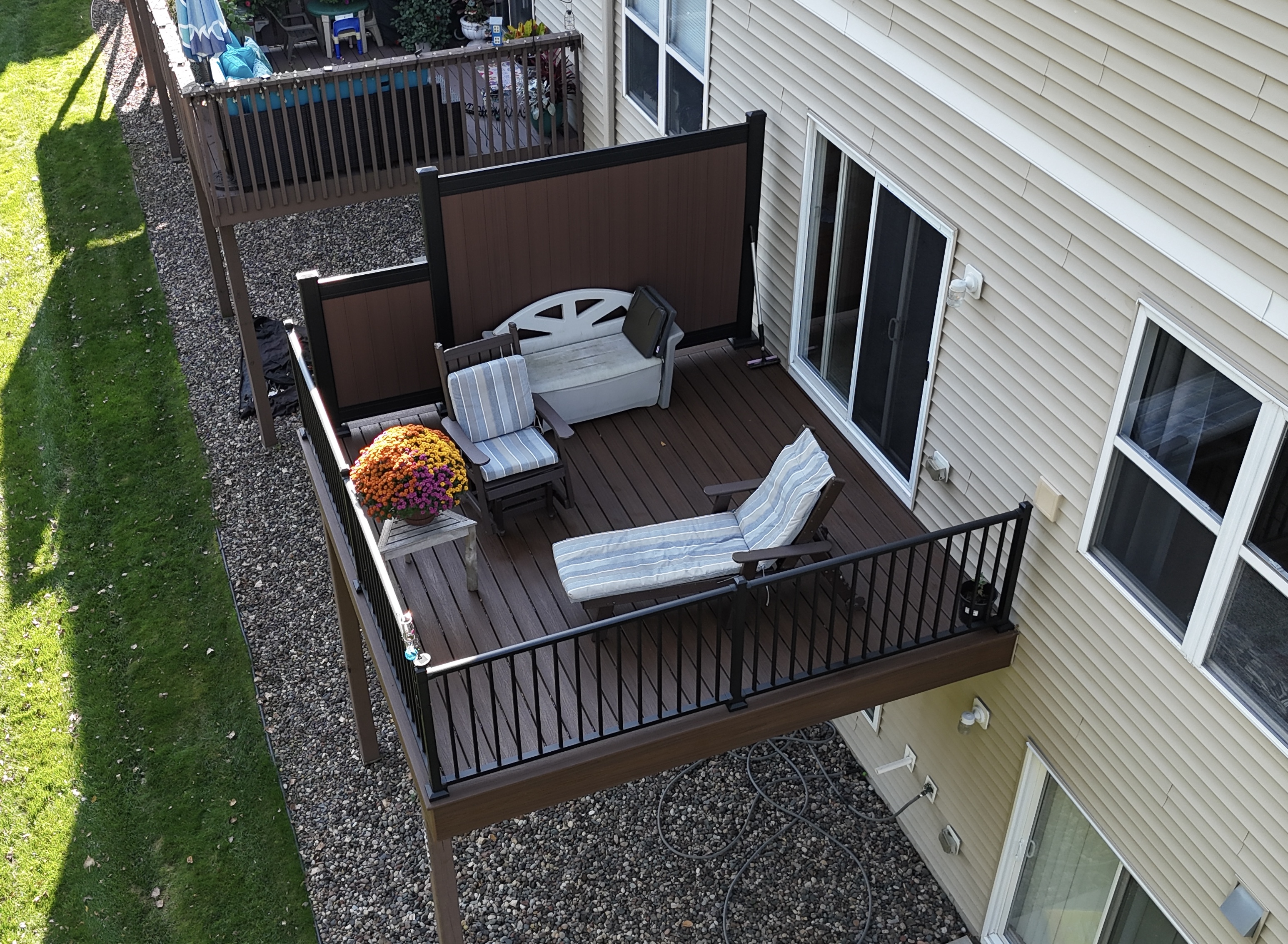 Wooden balcony with cushioned lounge chair, armchair, storage bench, and a small table holding a colorful flower pot.