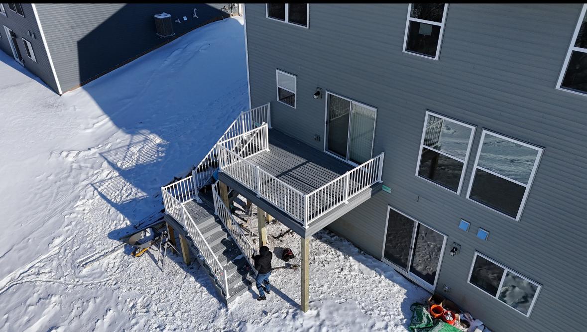 Raised deck with white railing and stairs attached to a gray two-story house, snowy ground and person working on the stairs.