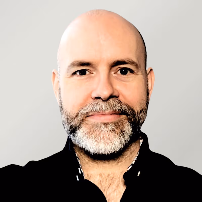 Close-up of a smiling bald man with a salt-and-pepper beard wearing a black shirt against a plain background.
