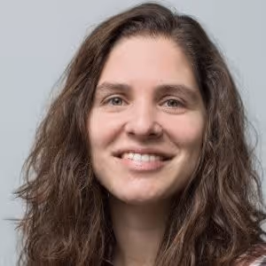 Woman with long curly brown hair smiling against a gray background.