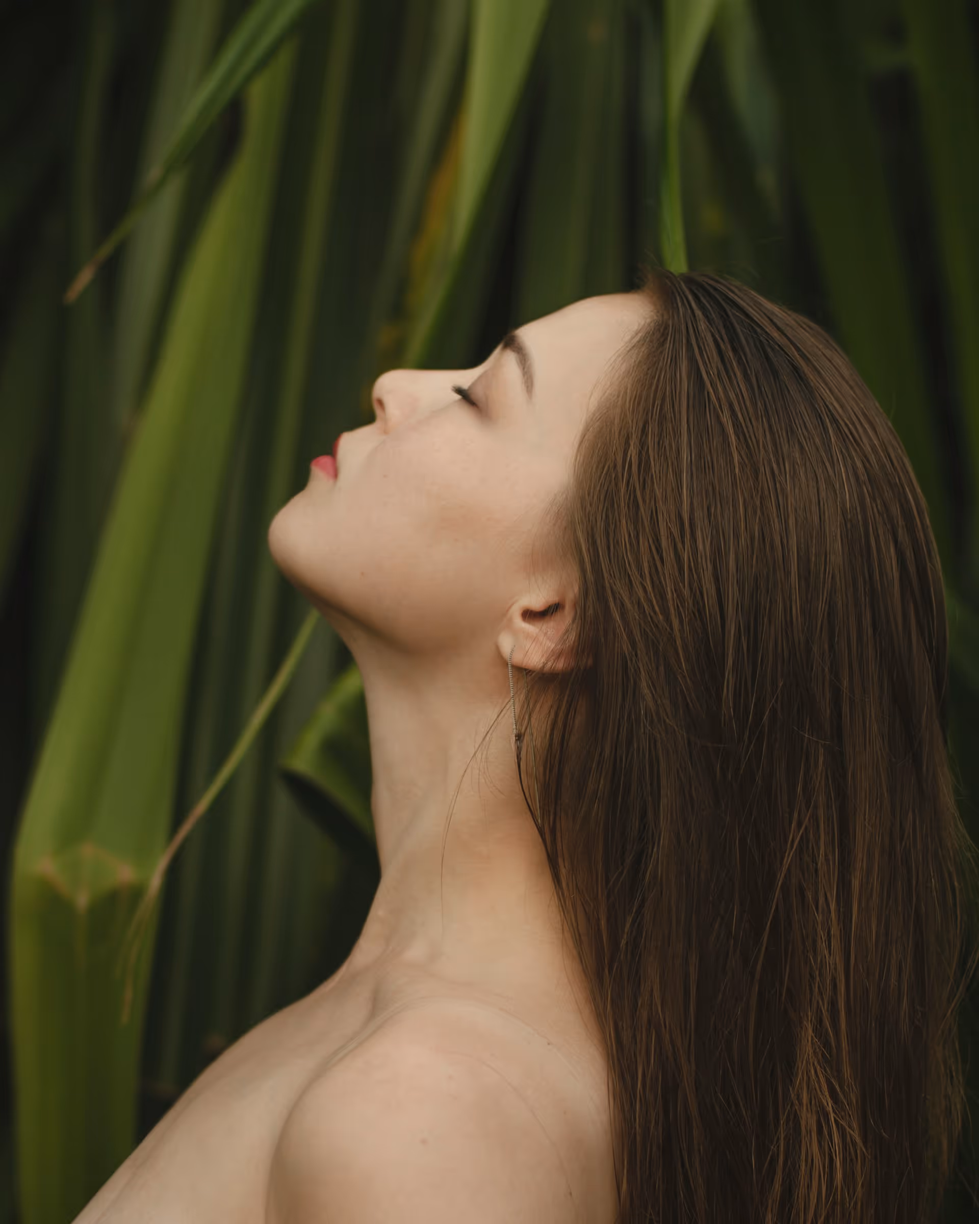 Profile of a woman with closed eyes and long brown hair against a background of green leaves.