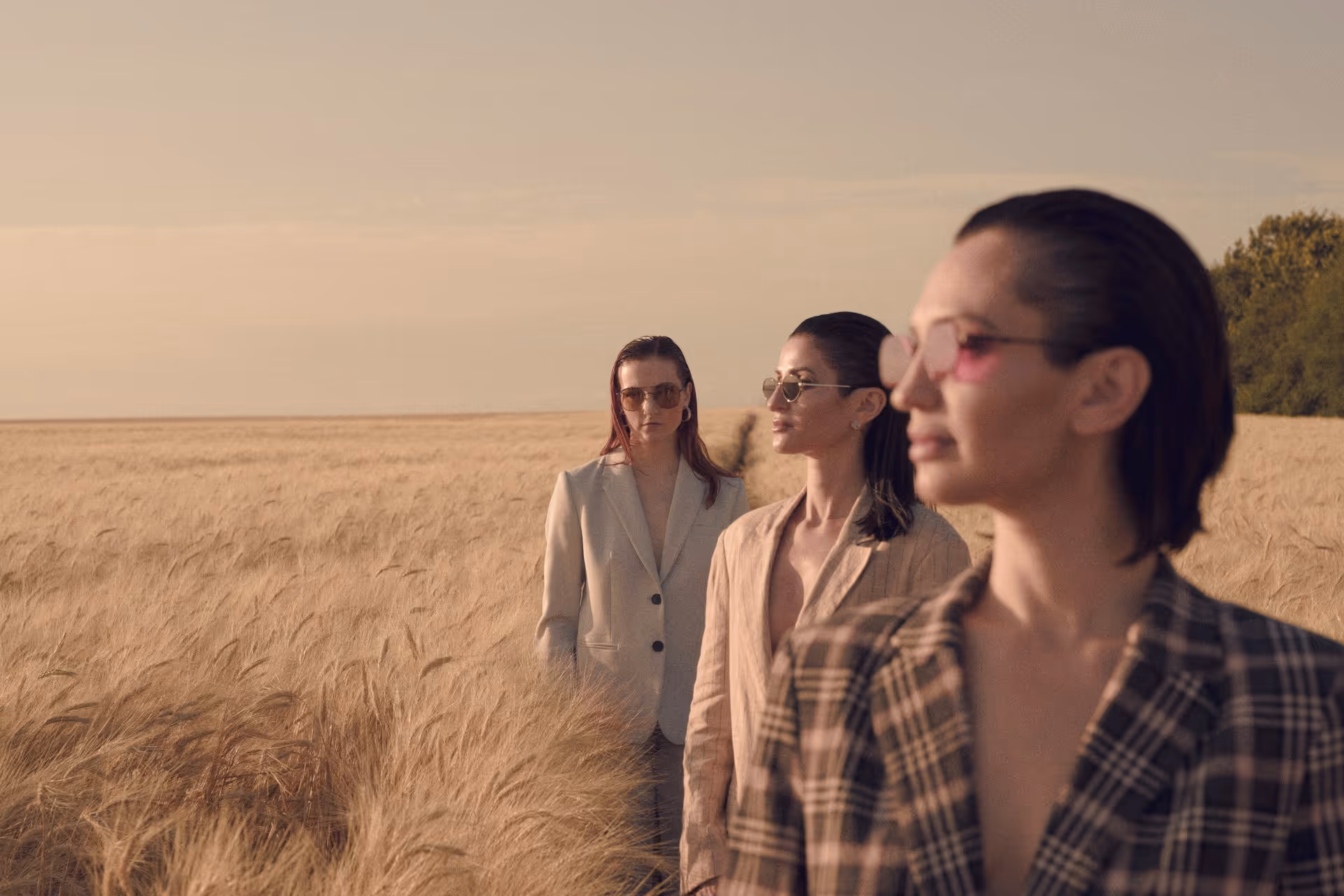 Three women wearing sunglasses and blazers standing in a golden wheat field during sunset.