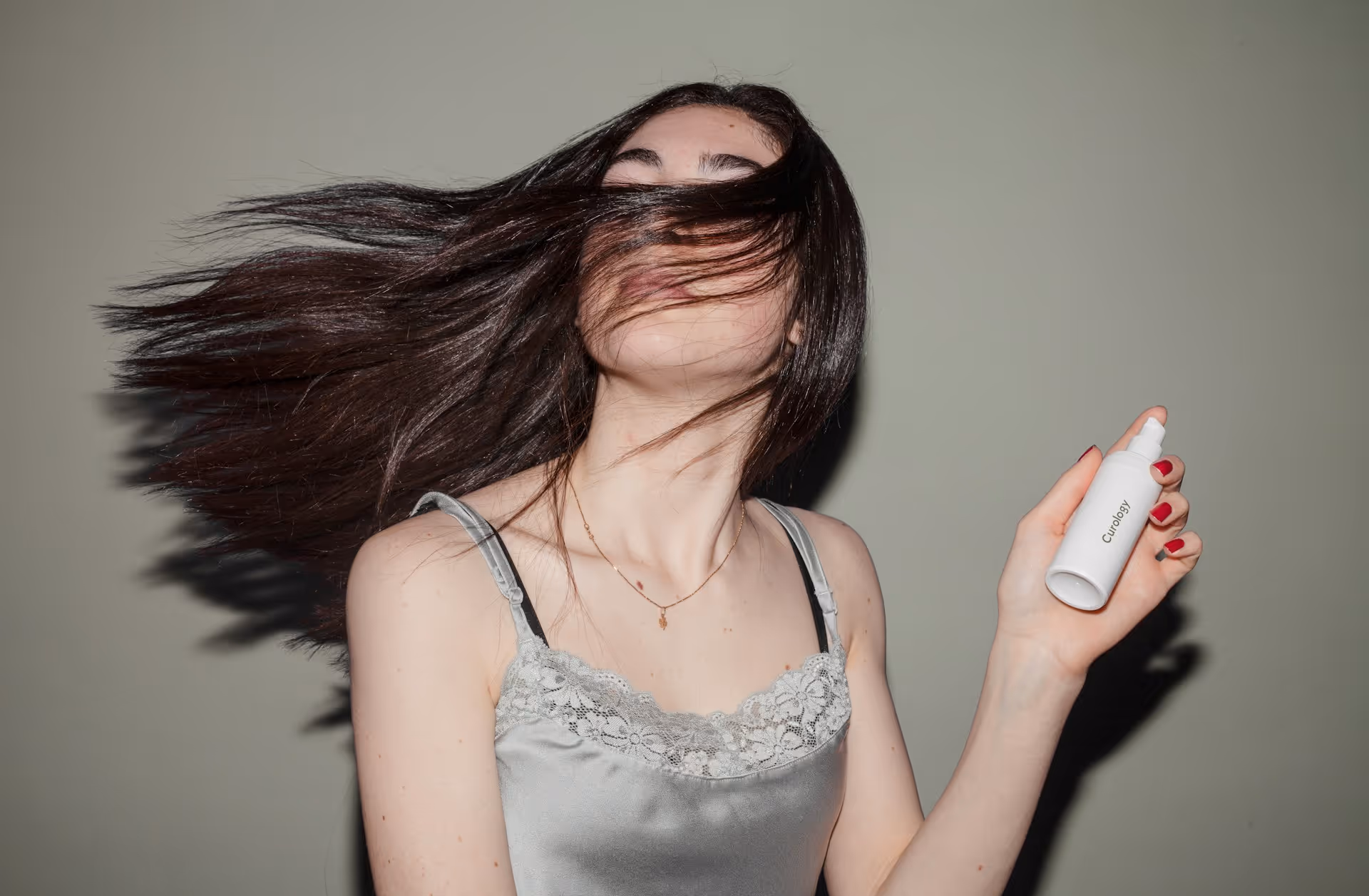 Woman in a silver lace-trimmed top with long dark hair flowing across her face holding a white Curology spray bottle.