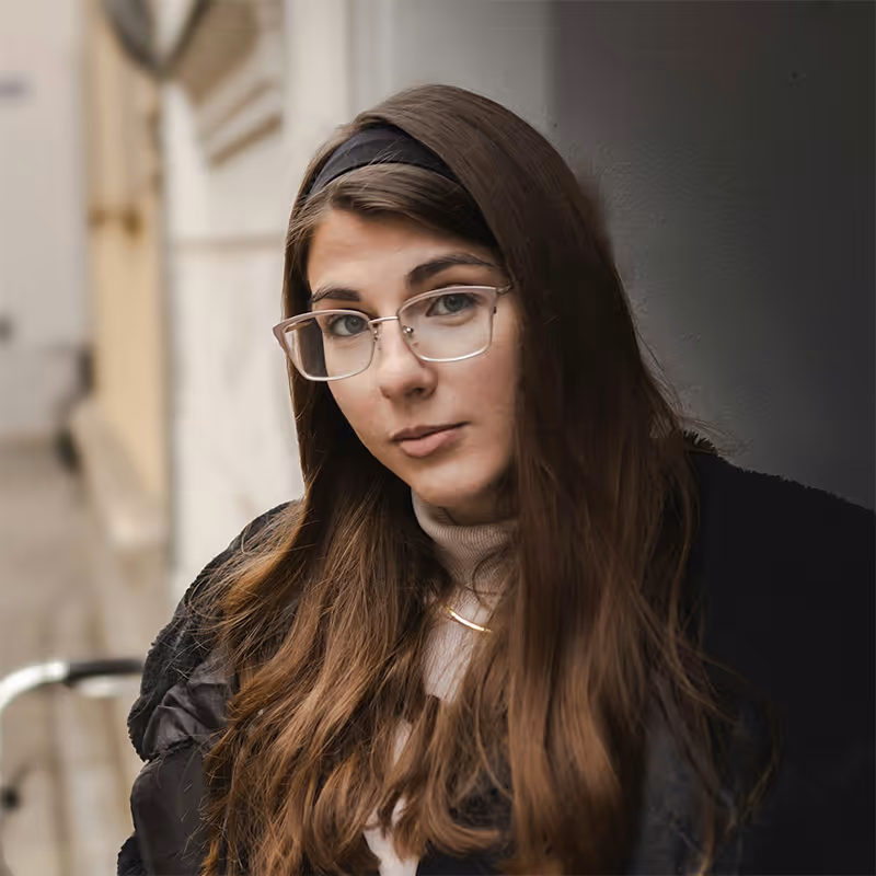 Young woman with long brown hair and glasses, wearing a black coat and light-colored turtleneck, leaning against a wall outdoors.