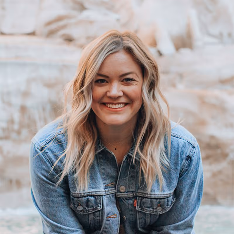 Smiling woman with long blonde hair wearing a denim jacket, in front of a softly blurred natural background.