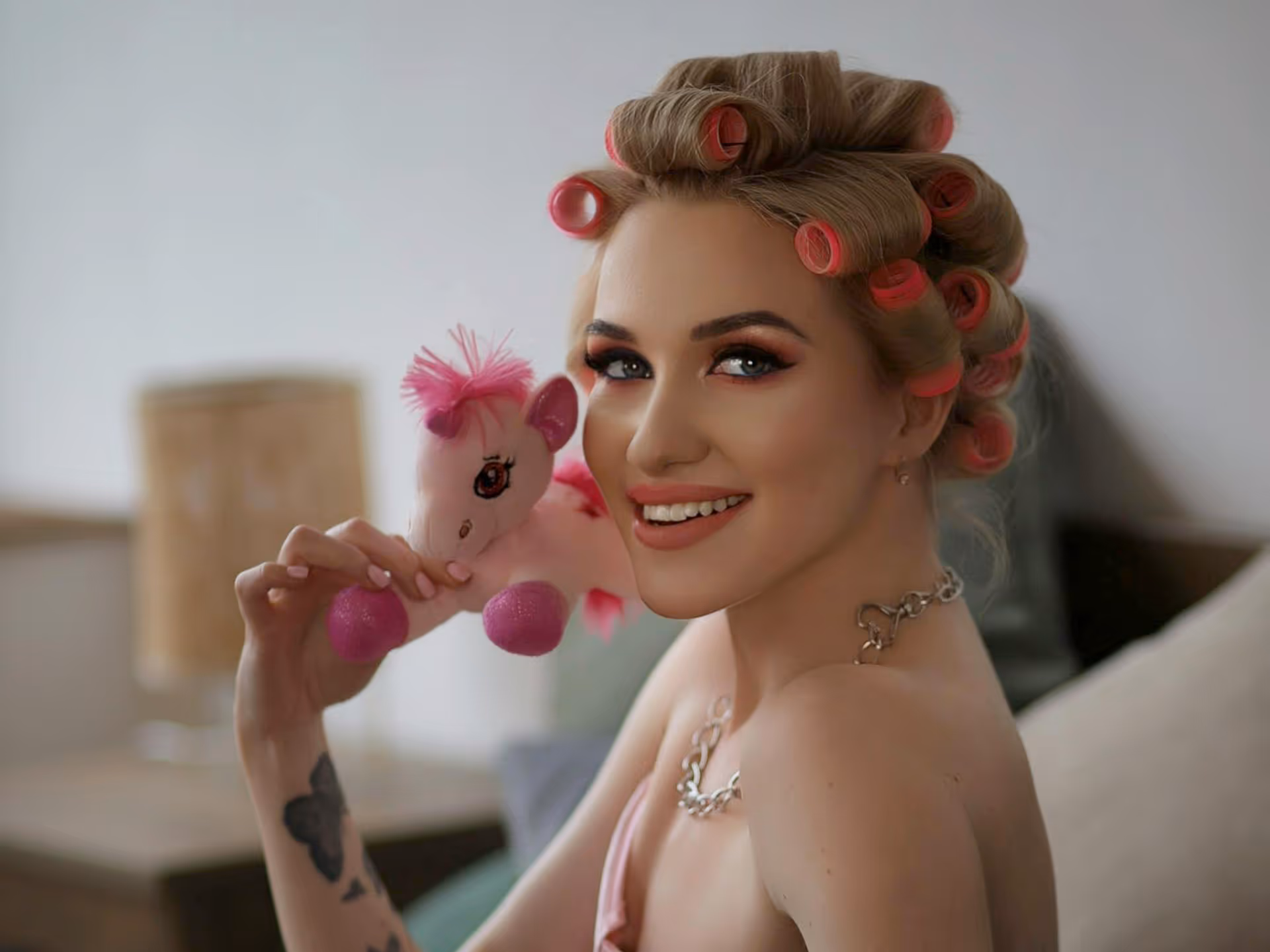 Smiling woman with pink hair rollers holding a pink stuffed unicorn toy.