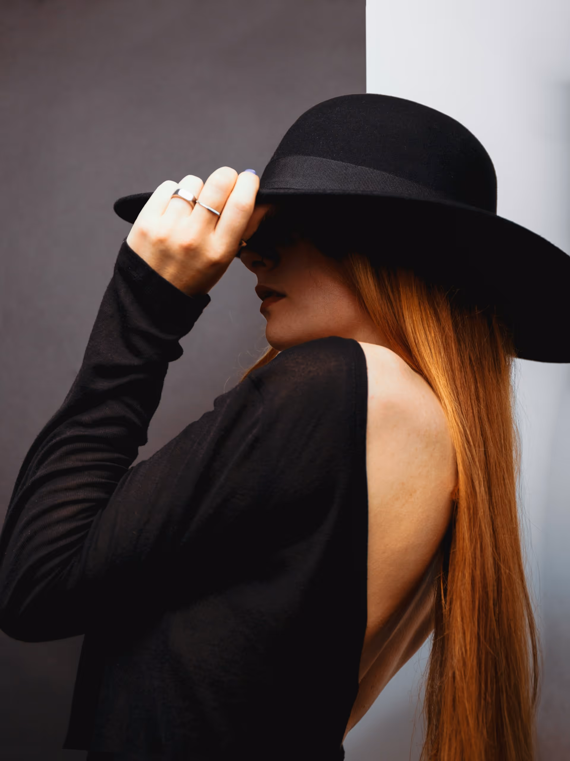 Woman with long red hair wearing a wide-brimmed black hat and a black backless top, holding the brim of her hat.