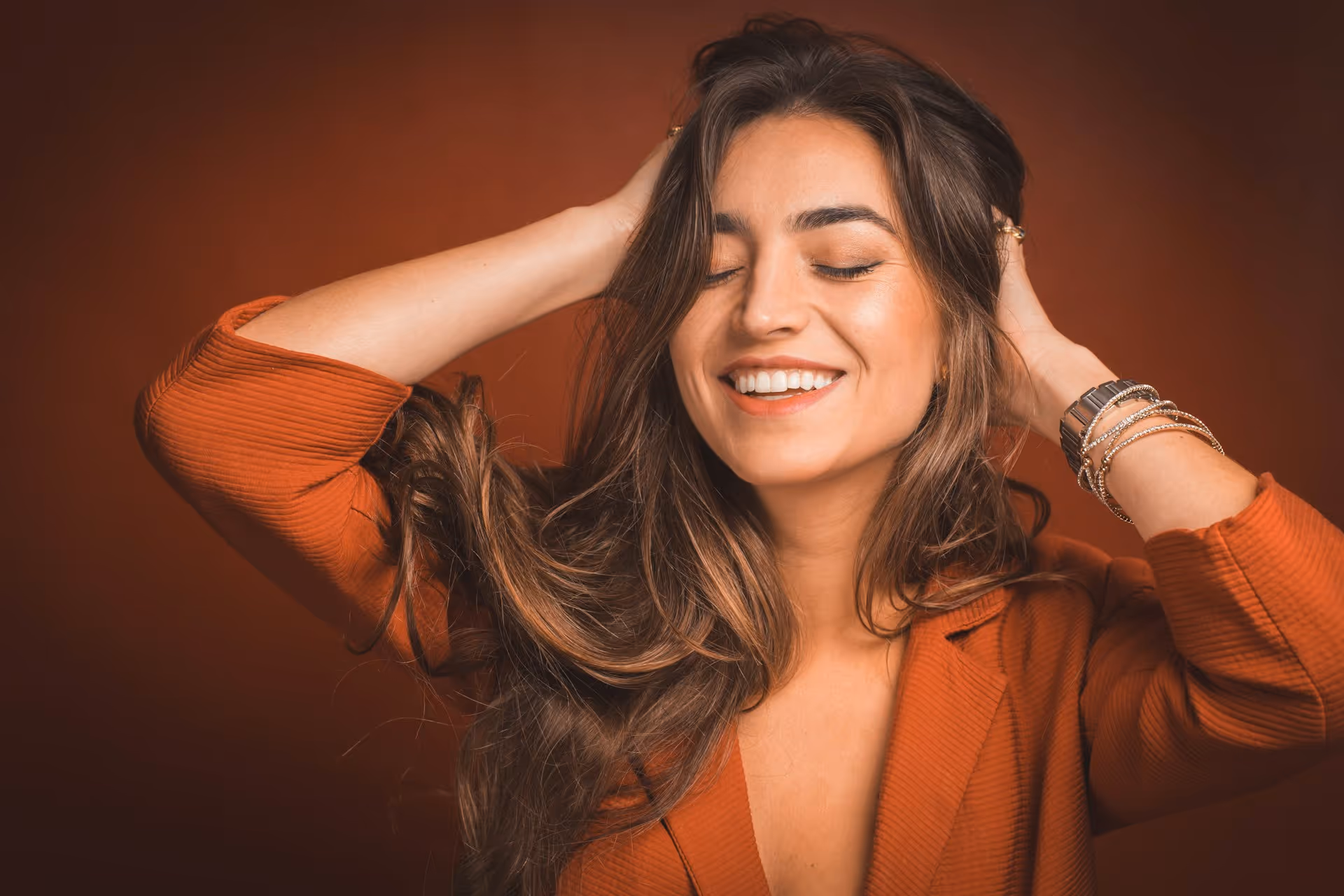 Smiling woman with long brown hair wearing a rust-colored jacket, with eyes closed and hands in her hair against a matching rust-colored background.