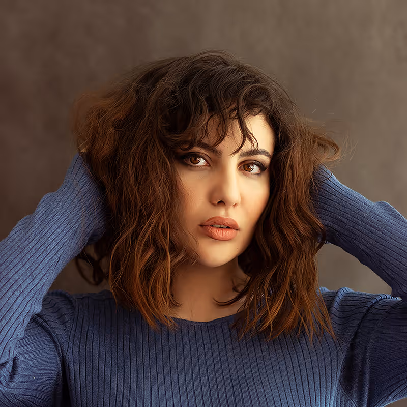 Woman with shoulder-length brown wavy hair wearing a blue ribbed sweater, posing with hands behind her head against a brown background.