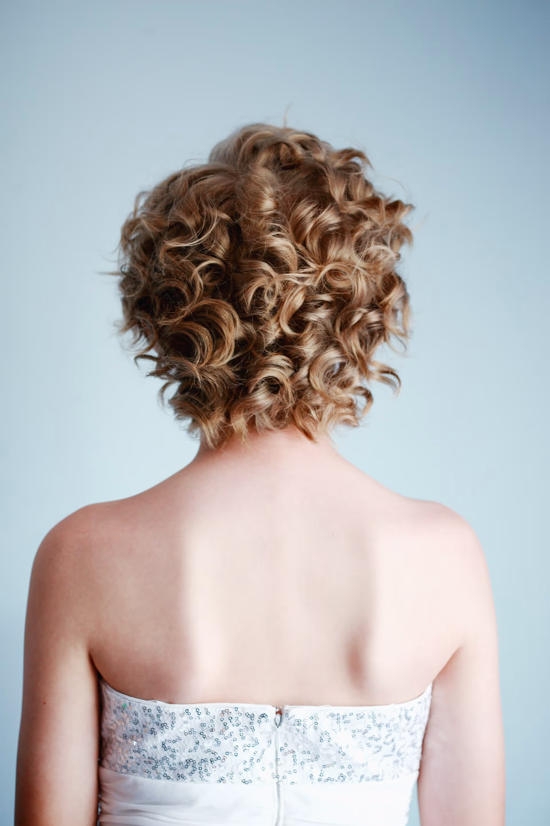 Person with short, voluminous curly hair wearing a strapless white sequined dress, shown from the back against a light blue background.