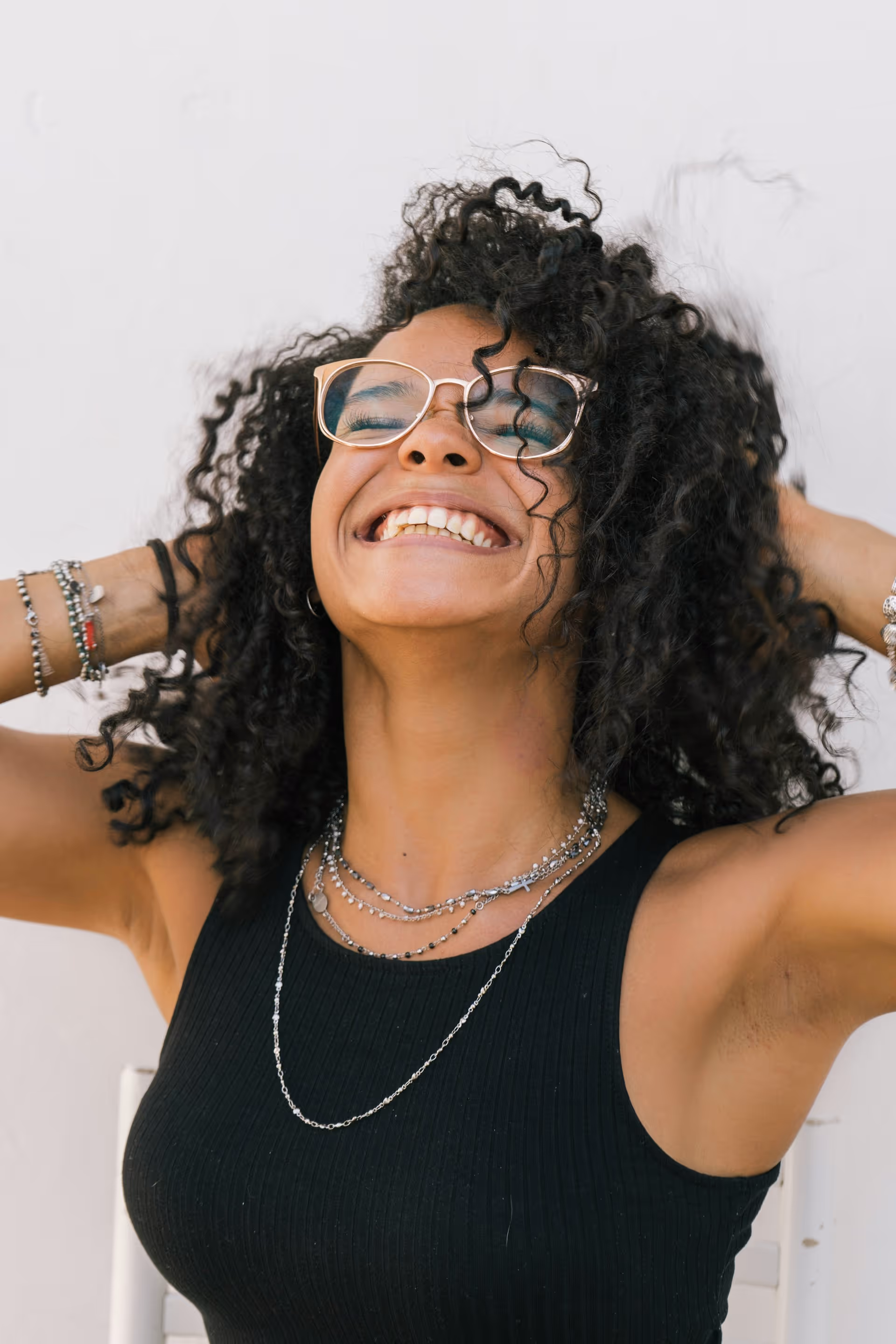Smiling woman with curly hair wearing glasses, a black sleeveless top, and layered silver necklaces and bracelets.