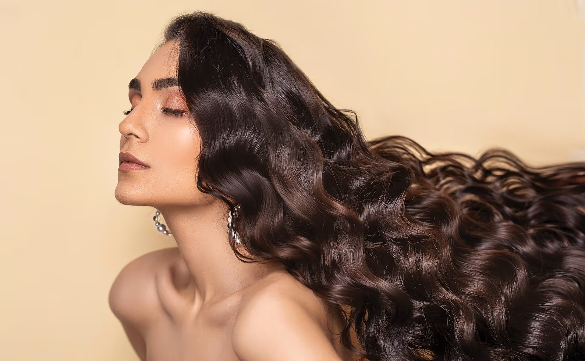 Profile of a woman with long, glossy, wavy dark brown hair and closed eyes against a beige background.