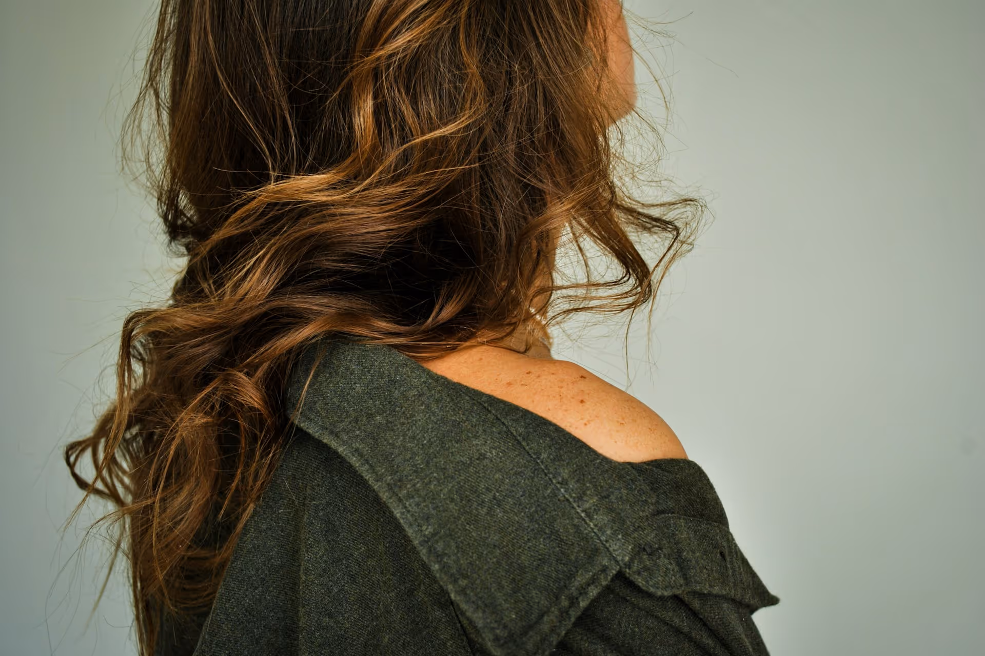 Close-up of a person with long, wavy brown hair wearing a dark green garment off one shoulder against a plain gray background.