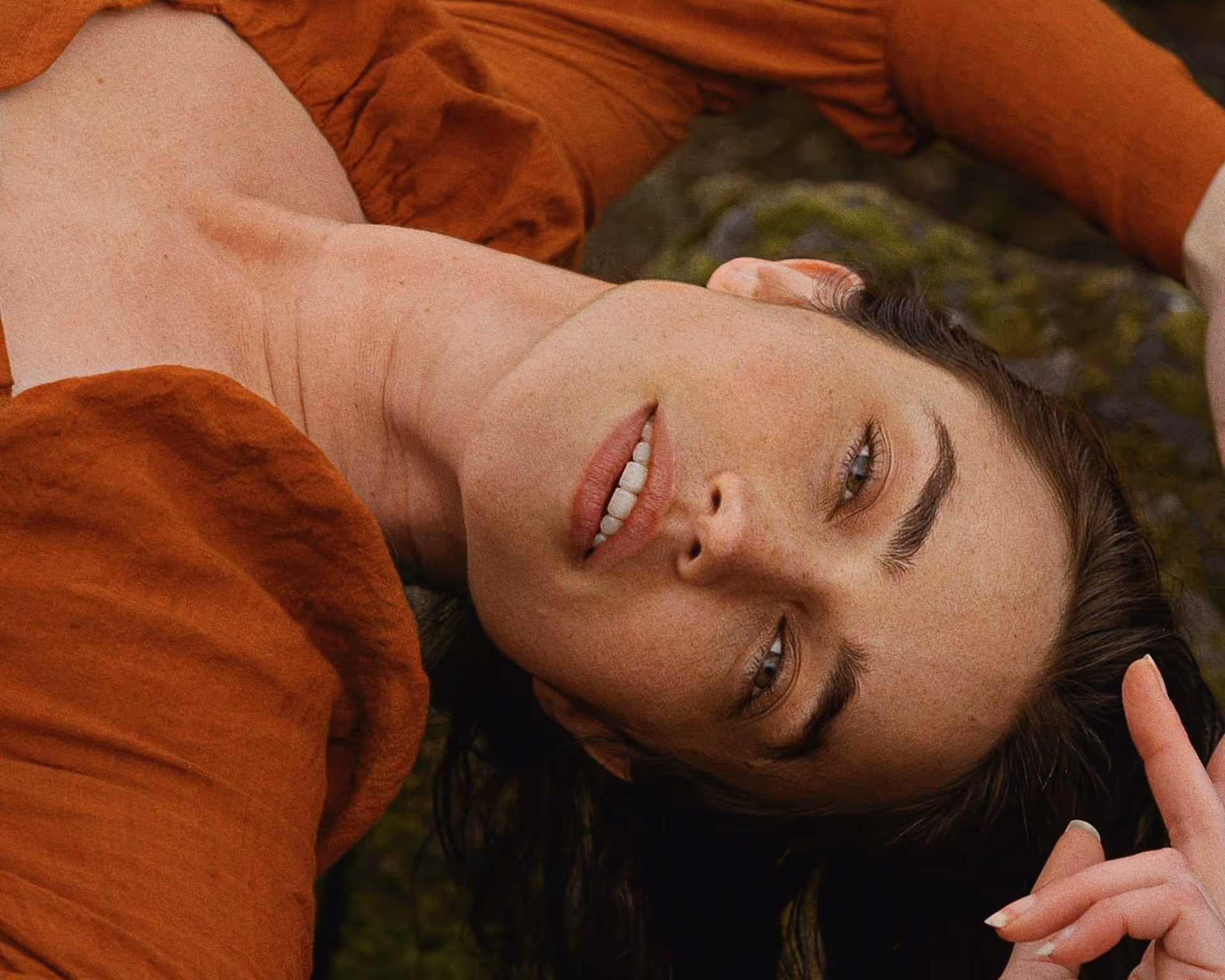 Close-up of a woman with light skin and green eyes lying down, wearing a rust-colored top.