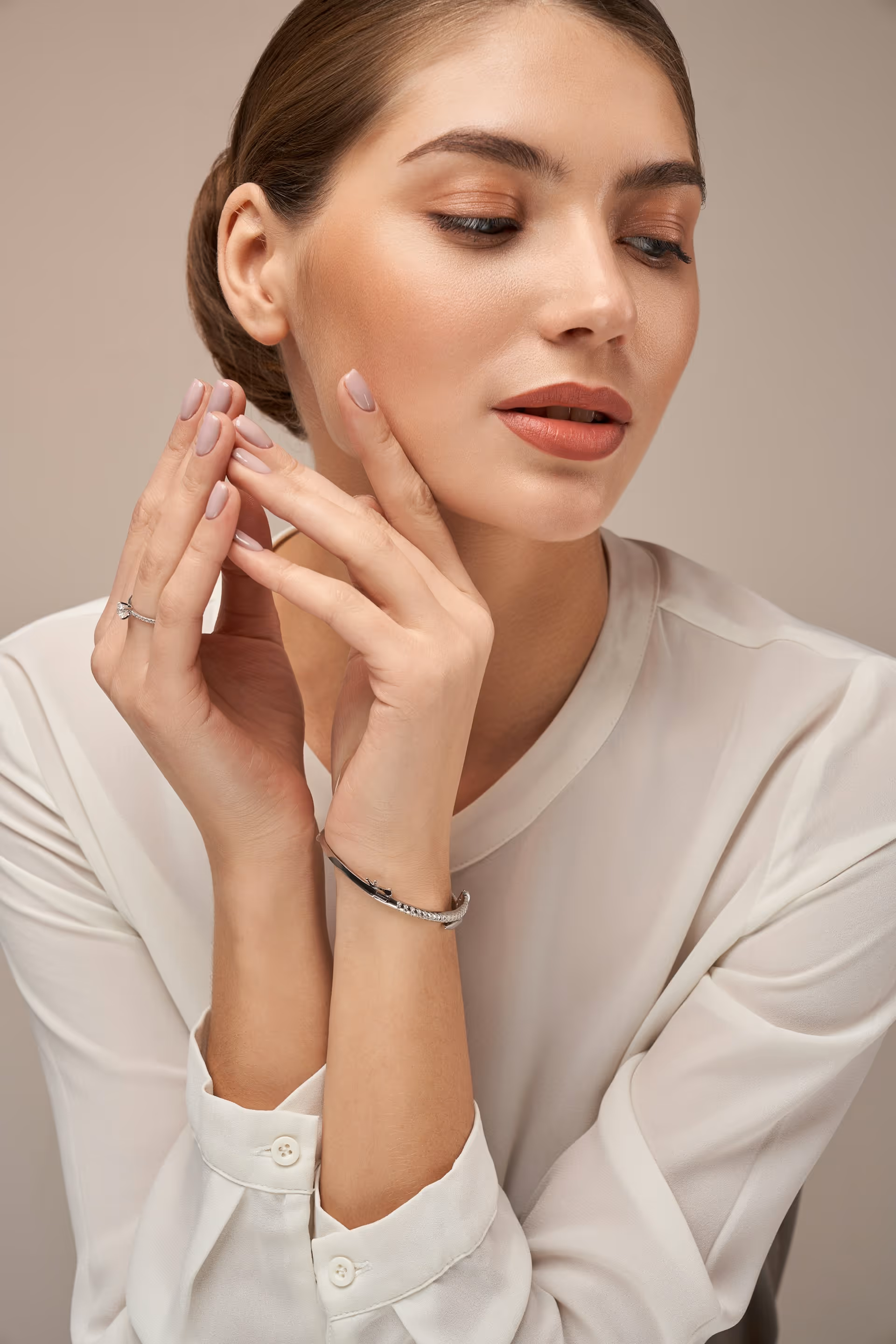 Woman with neatly styled hair wearing a white blouse, showcasing a diamond ring and a bracelet with her hands near her face.