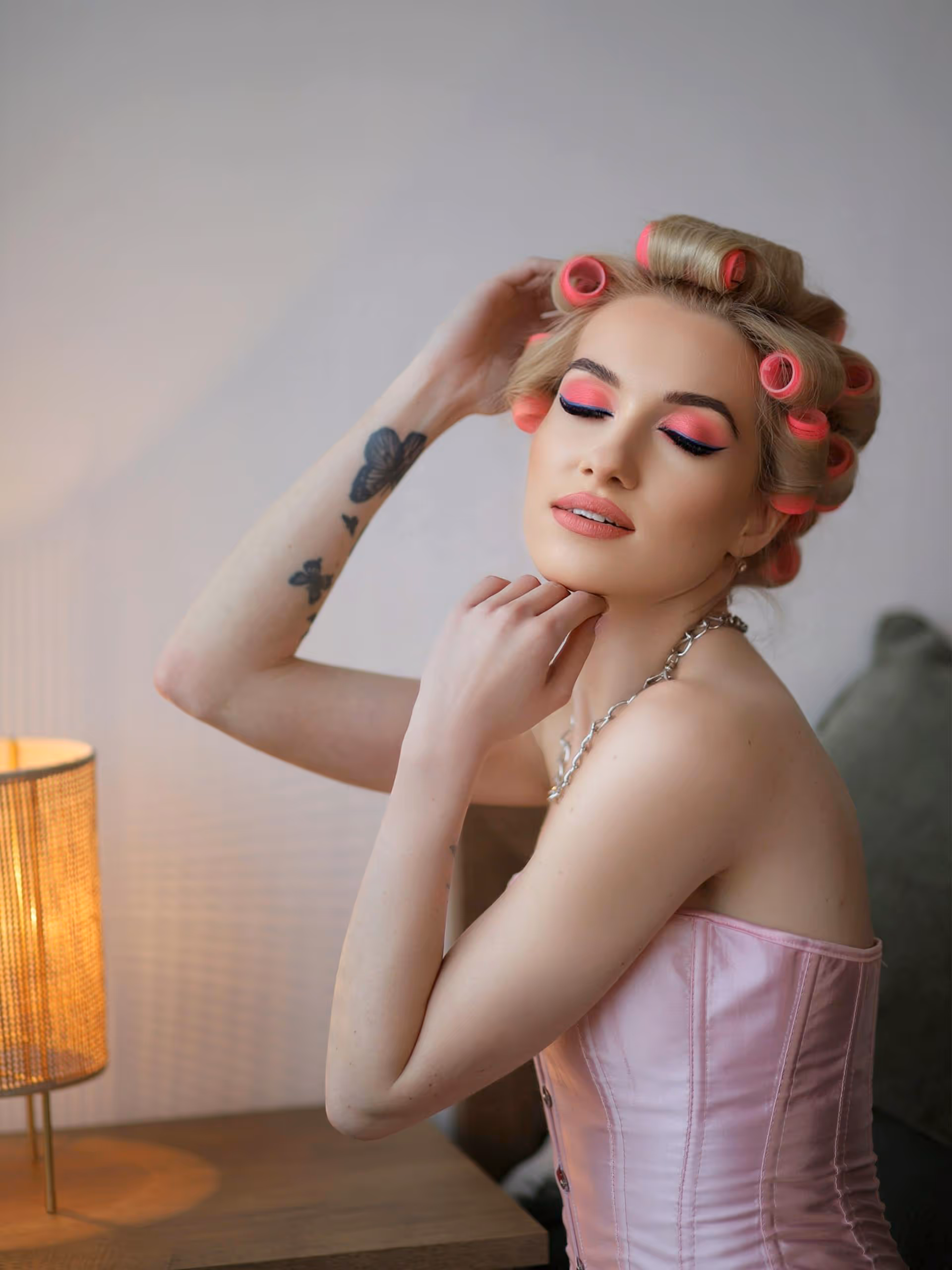 Woman with closed eyes wearing pink eye makeup and pink hair rollers, dressed in a light pink corset top, sitting beside a wooden table with a lit lamp.