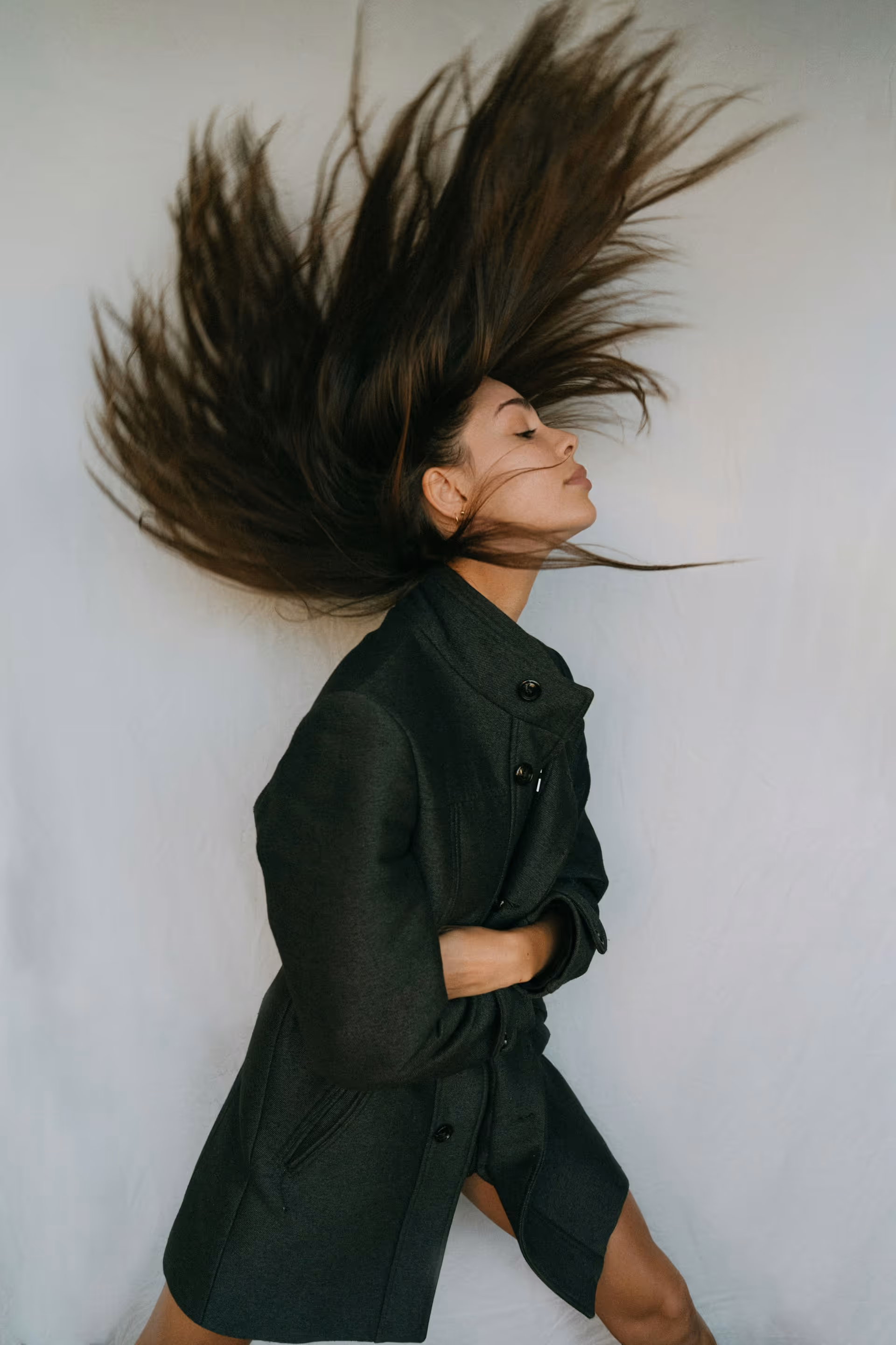 Woman with long brown hair flipping it back while wearing a dark grey coat against a plain backdrop.
