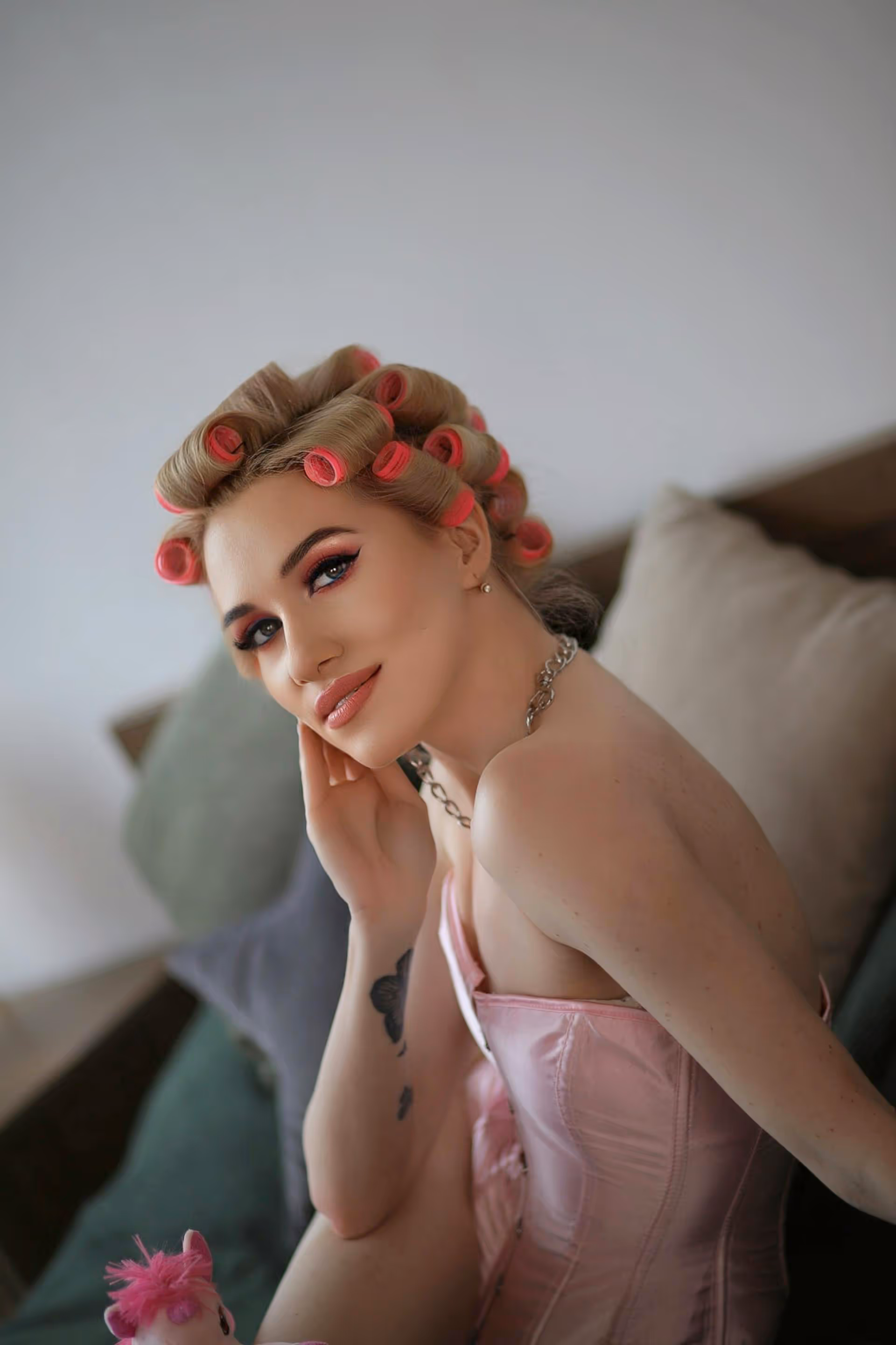 Woman with blonde hair in pink curlers wearing a pink satin corset smiling while sitting on a couch with cushions.