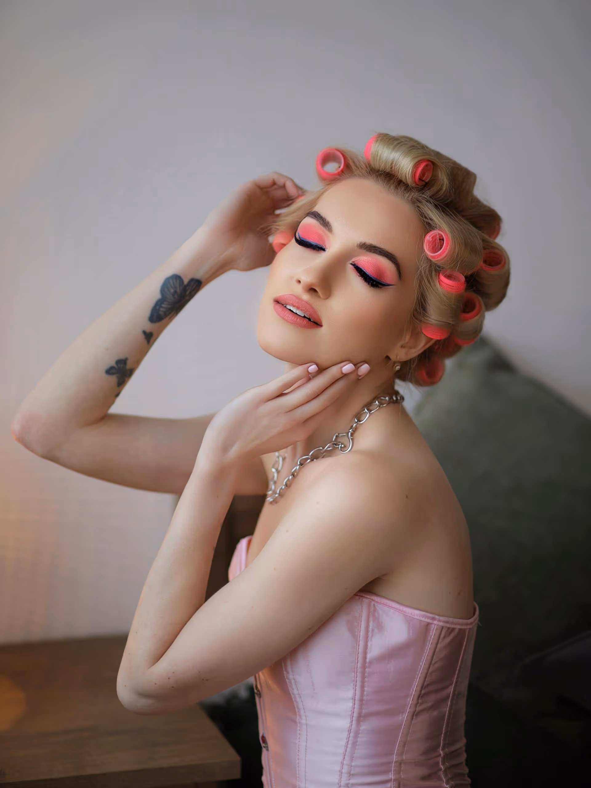Woman with blonde hair in pink curlers posing with closed eyes and pink eyeshadow, wearing a pink satin corset and a silver chain necklace.