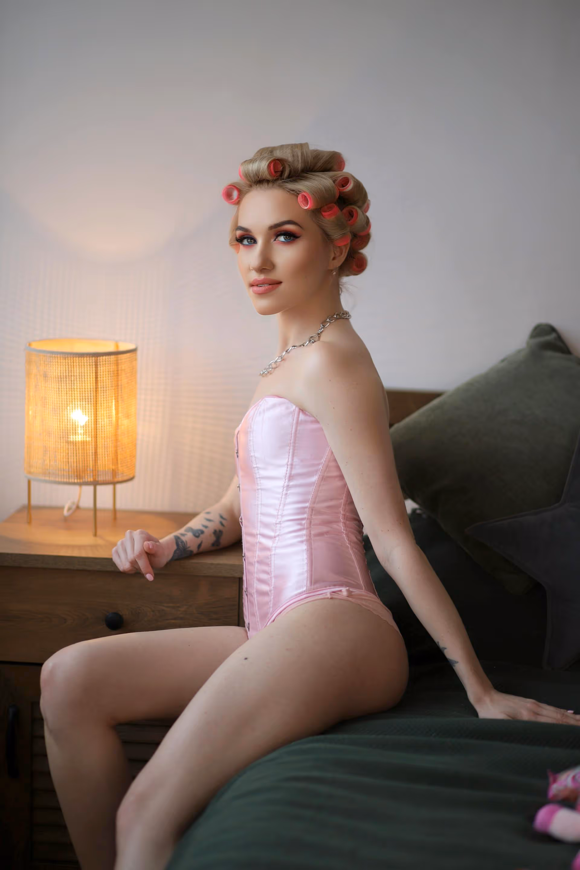 Woman with hair curlers wearing a pink corset sitting on a bed next to a lit bedside lamp.