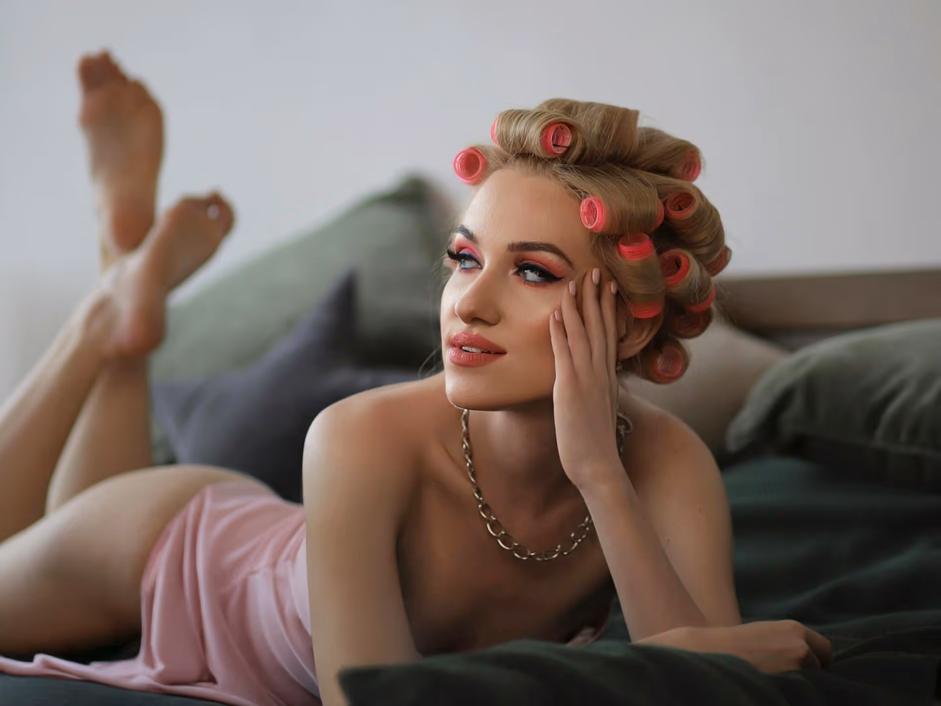 Woman lying on a bed with pink hair rollers, wearing a pink dress and a chain necklace, resting her head on her hand.