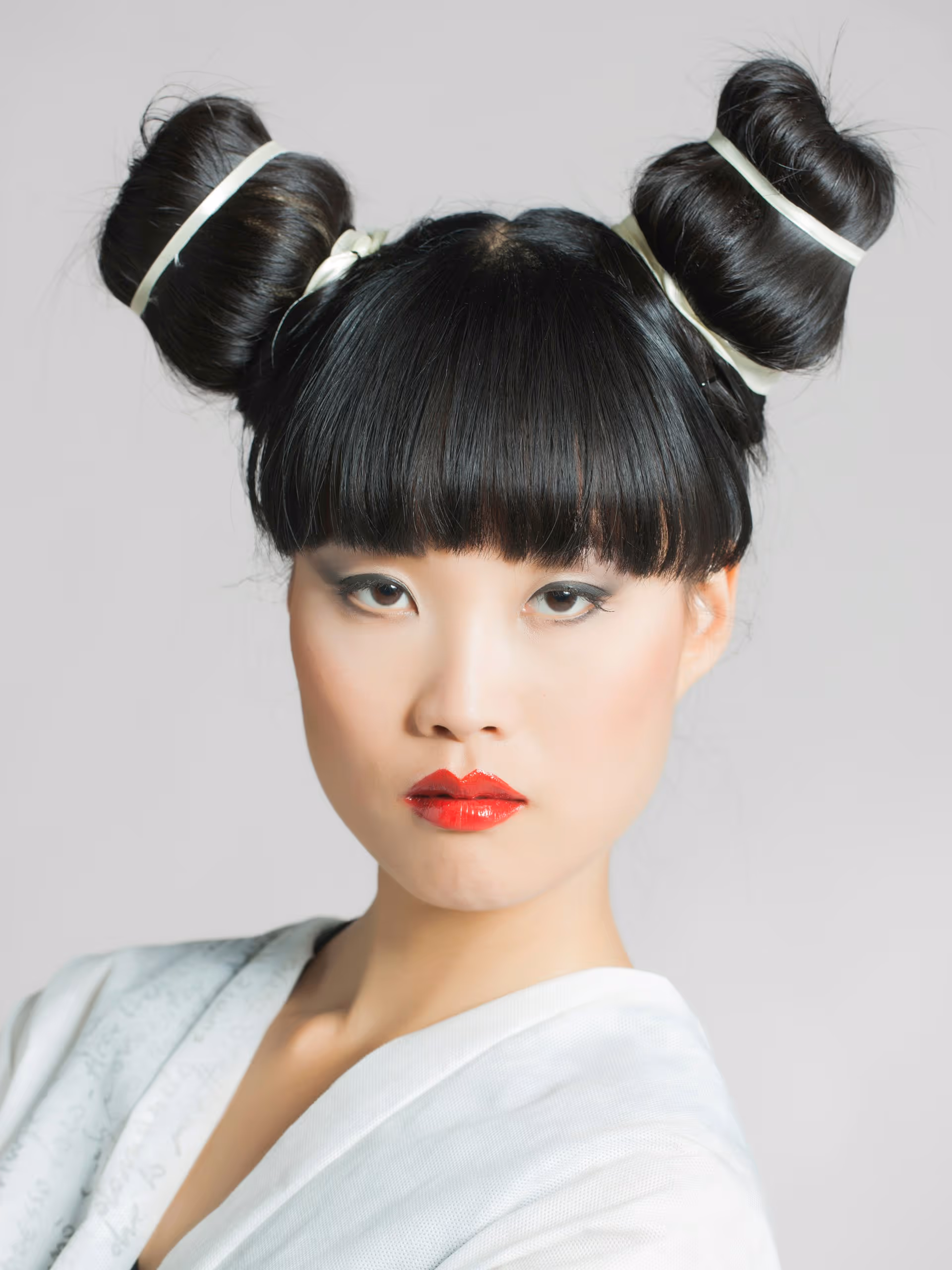 Portrait of an Asian woman with black hair styled in two buns, red lipstick, and pale makeup wearing a white garment.